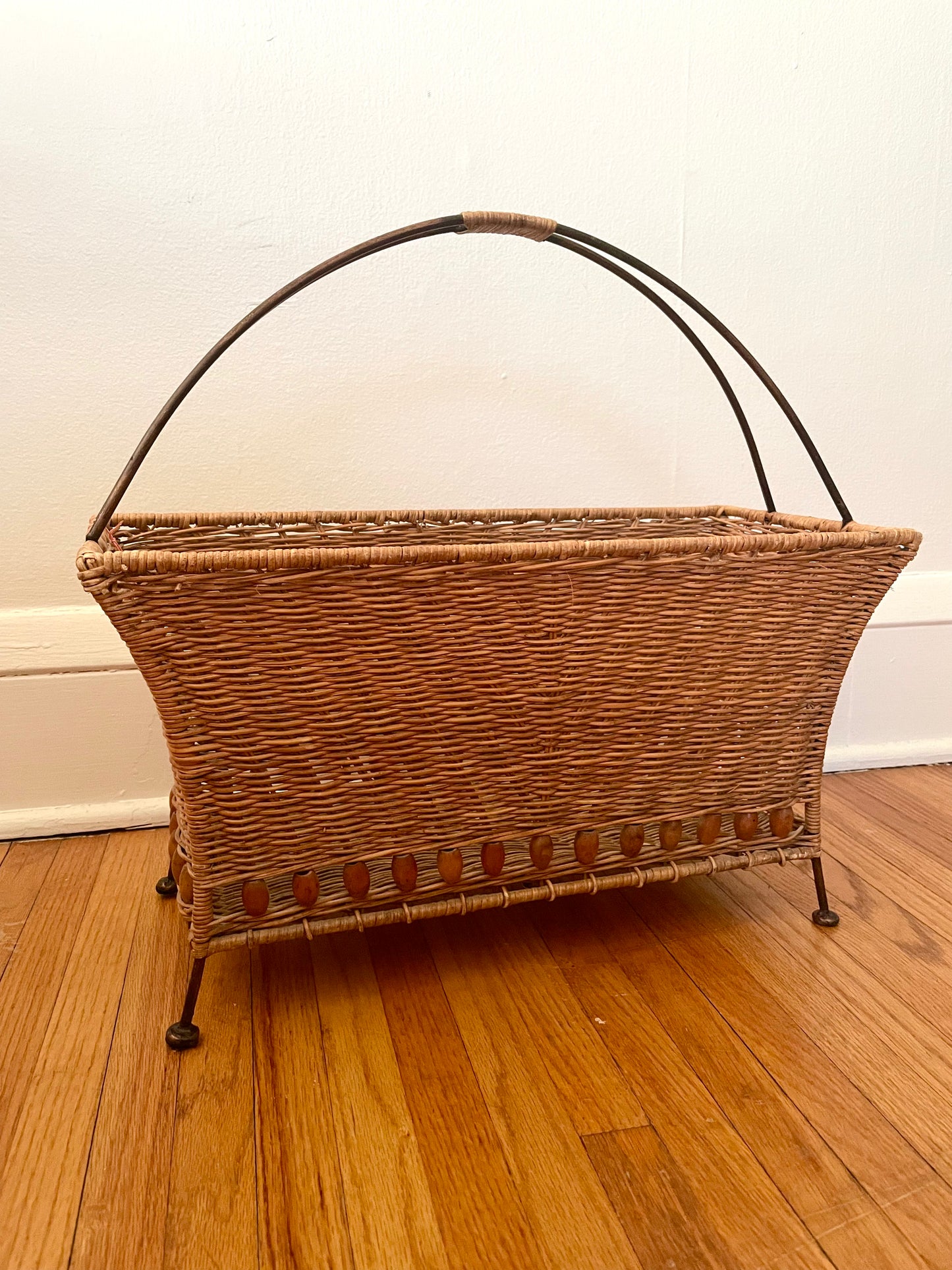 Wicker magazine basket with rattan-wrapped curved metal handle and metal feet, sitting on a wooden floor against a white wall.