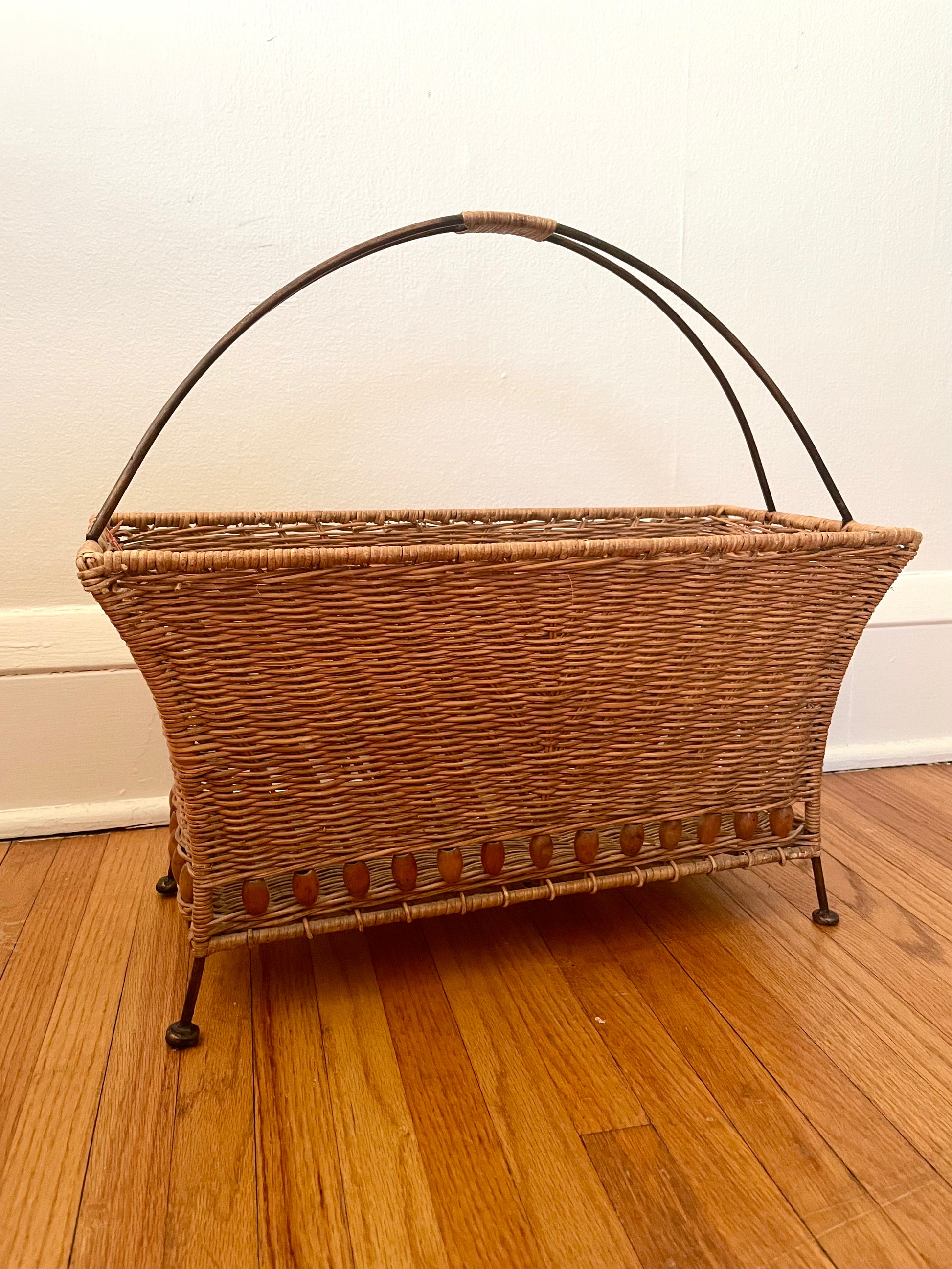Wicker magazine basket with rattan-wrapped curved metal handle and metal feet, sitting on a wooden floor against a white wall.