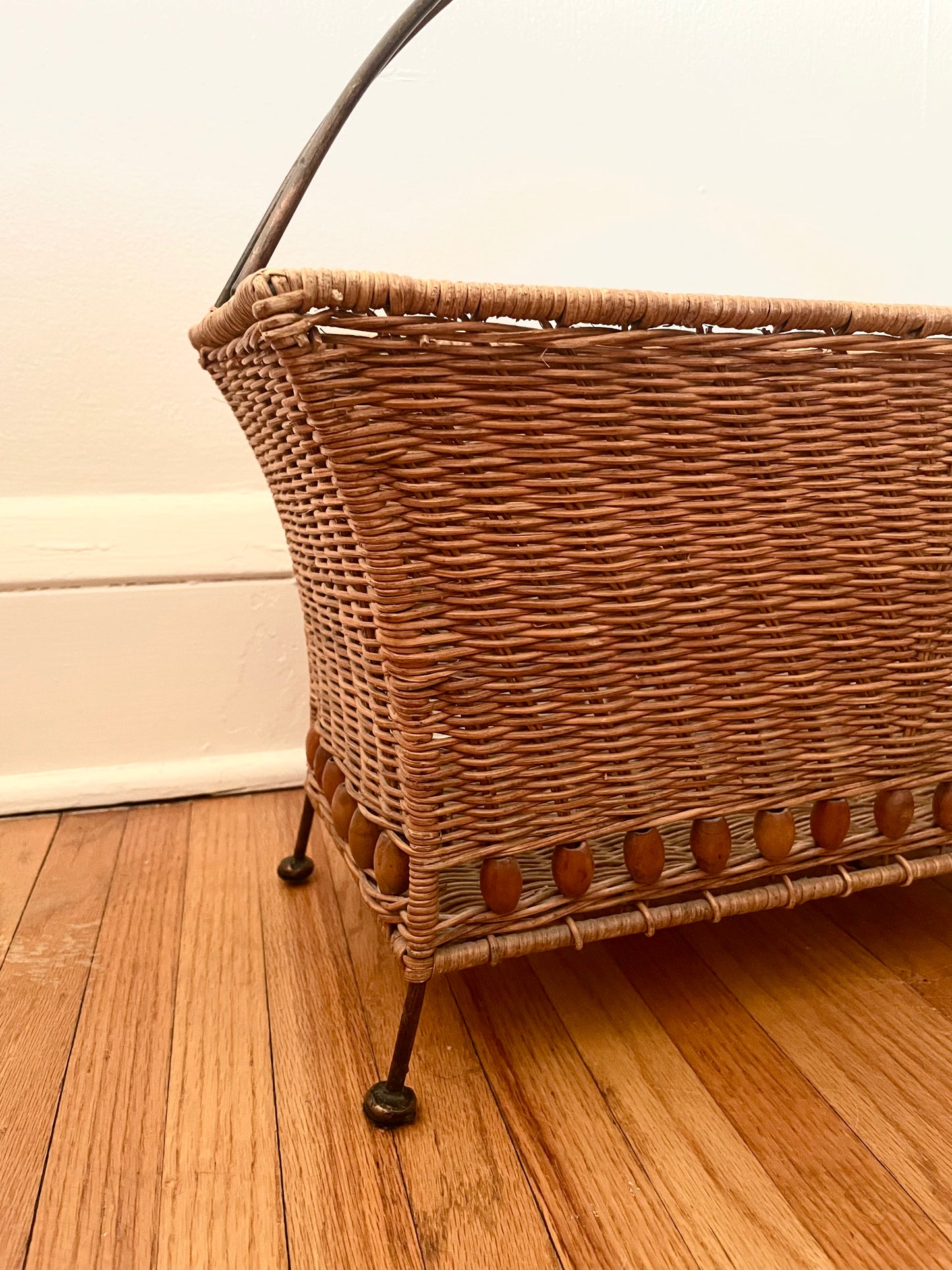 Partial view of a wicker magazine basket with rattan-wrapped curved metal handle and metal feet, sitting on a wooden floor against a white wall.