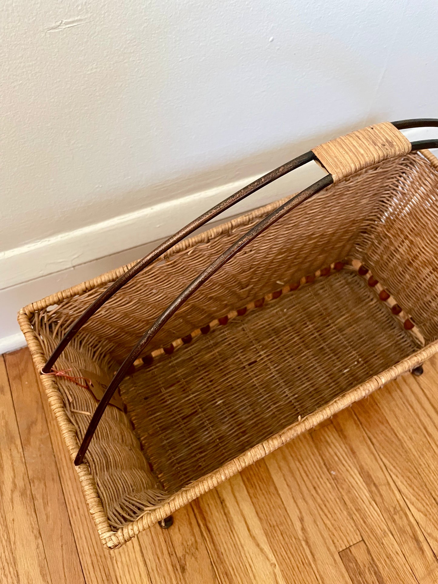Wicker magazine basket on a wooden floor against a white wall.