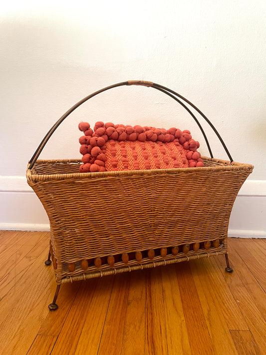 Wicker magazine basket with rattan-wrapped curved metal handle, metal feet, and red textured pillow inside, sitting on a wooden floor against a white wall.