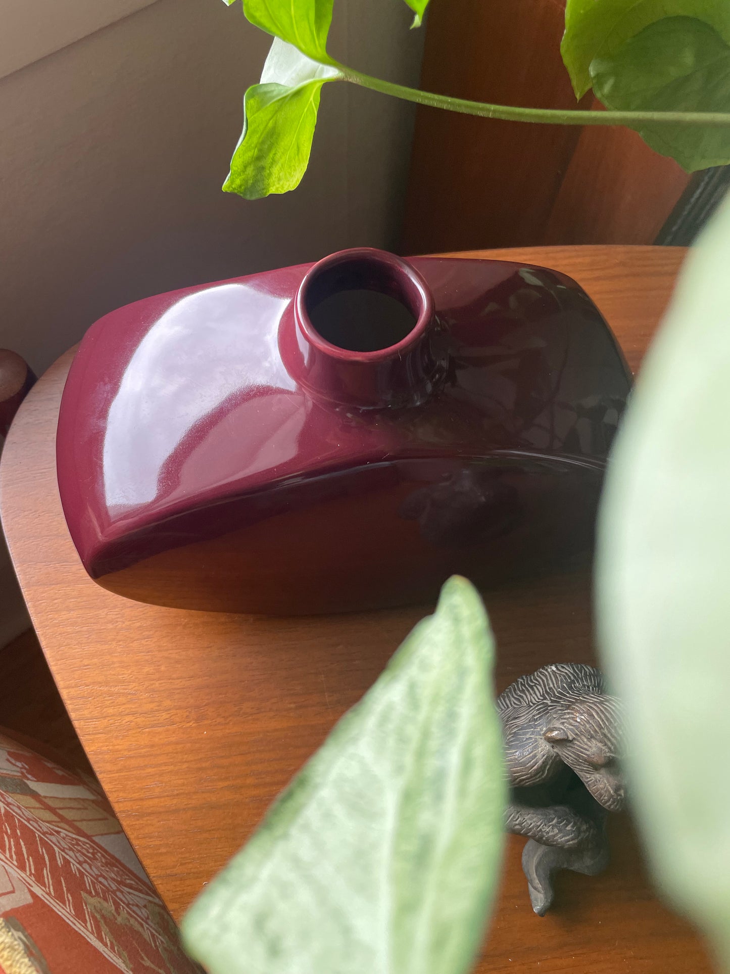 A glossy dark red ceramic vase with a small opening, displayed on a wooden table with houseplant leaves in the foreground.