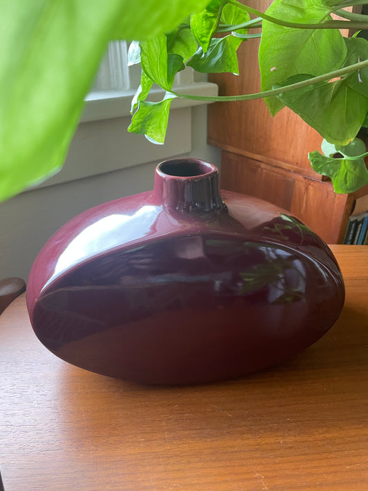 A glossy, rounded ceramic vase in a deep burgundy or maroon glaze, placed on a wooden table with a green houseplant in the foreground and a wooden cabinet in the background.