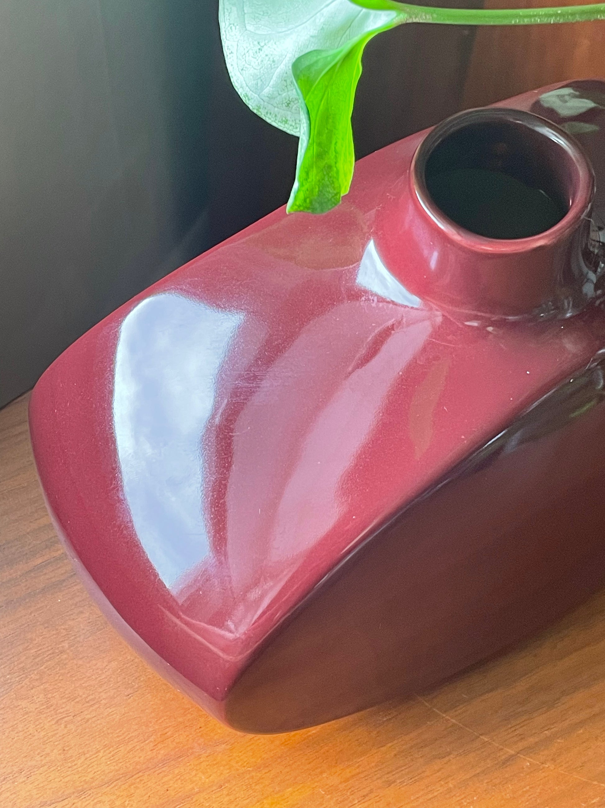 A glossy dark red ceramic vase with a small opening at the top, resting on a wooden surface with a green plant leaf visible above it.