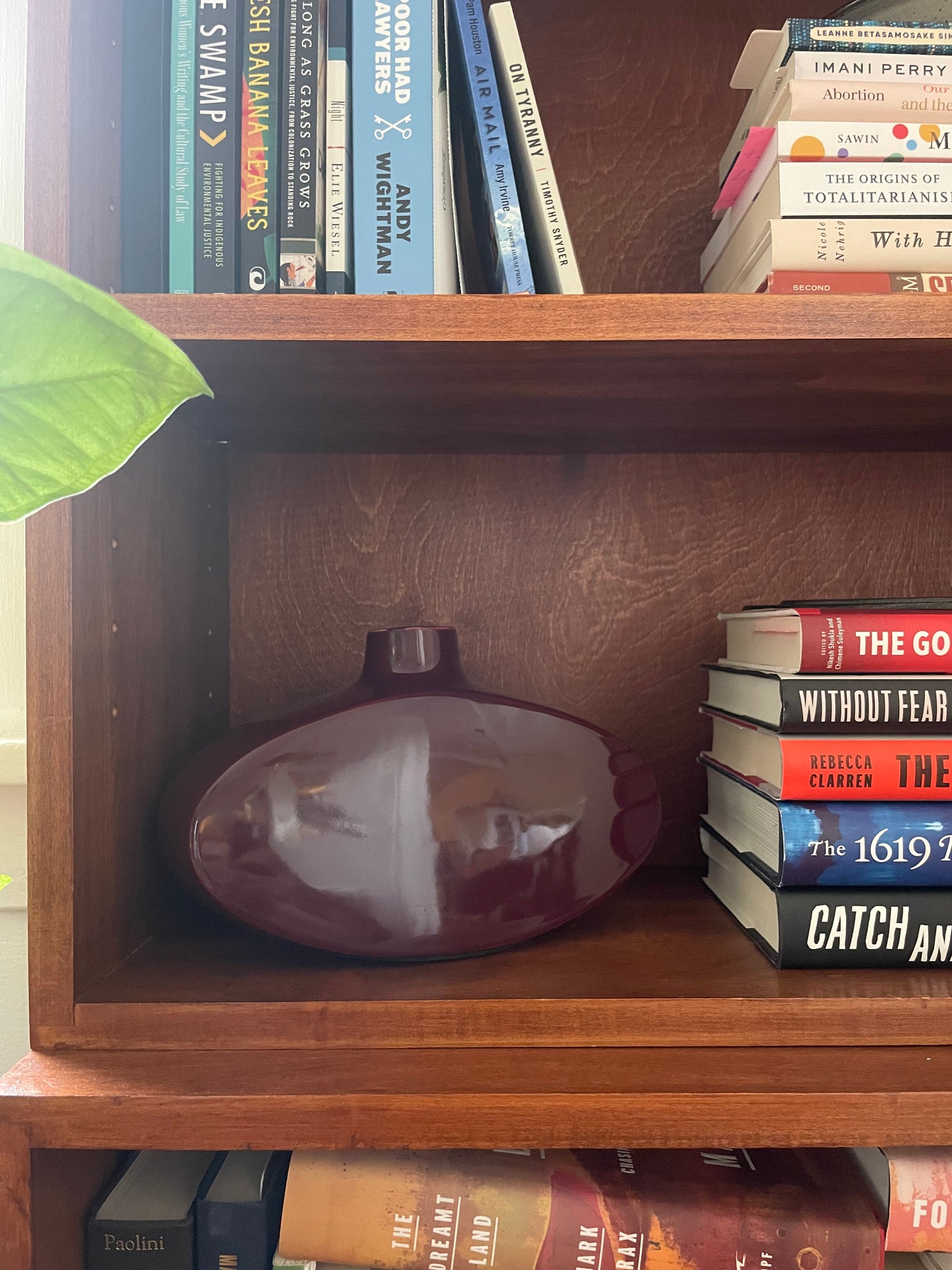 A glossy, rounded dark red ceramic vase displayed on a wooden bookshelf next to a stack of books.