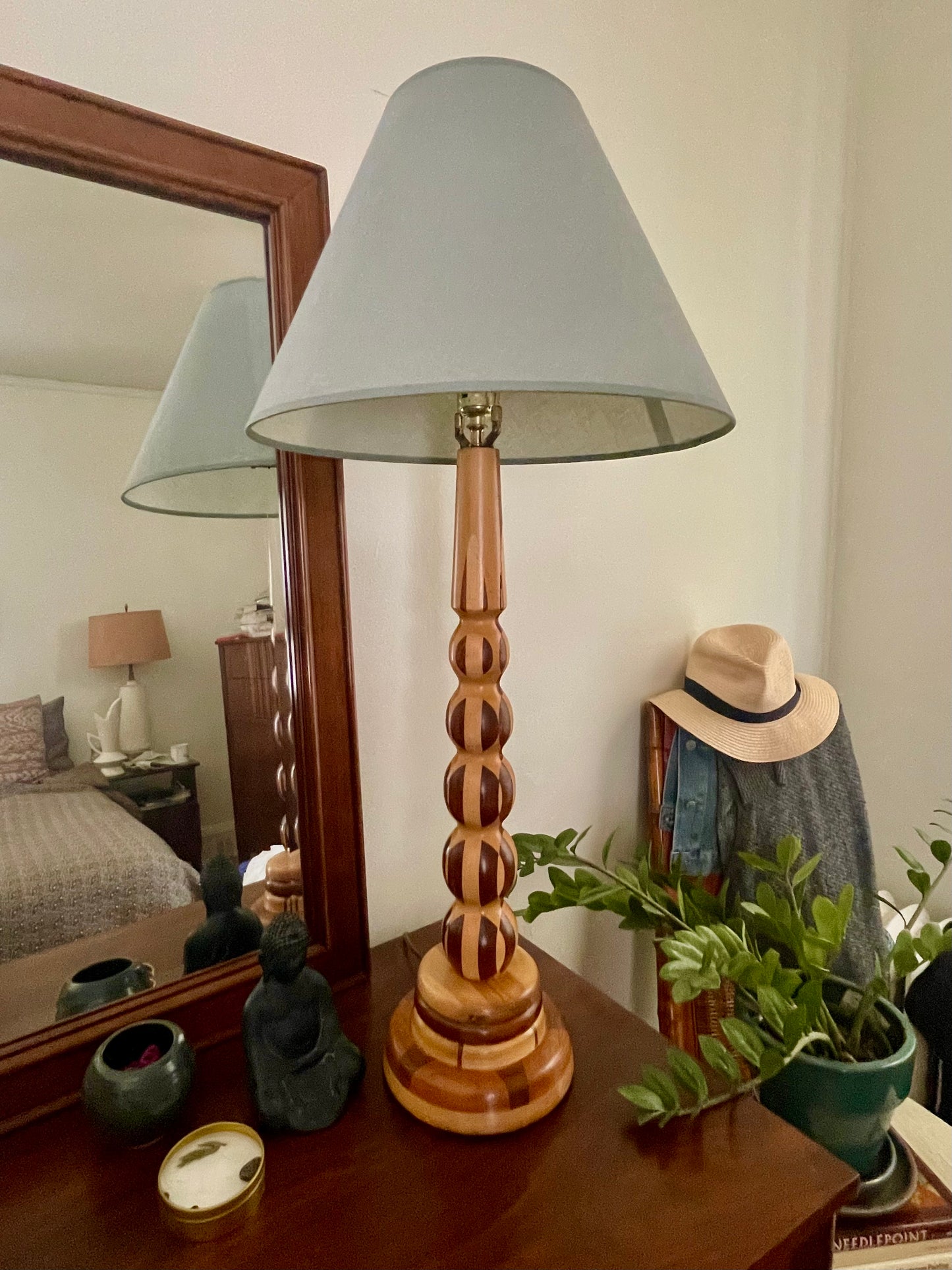 A wooden table lamp with a light blue-gray tapered linen shade and a turned parquetry wooden base, styled on a dark wood dresser in a bedroom setting with a mirror, hat, and plant.