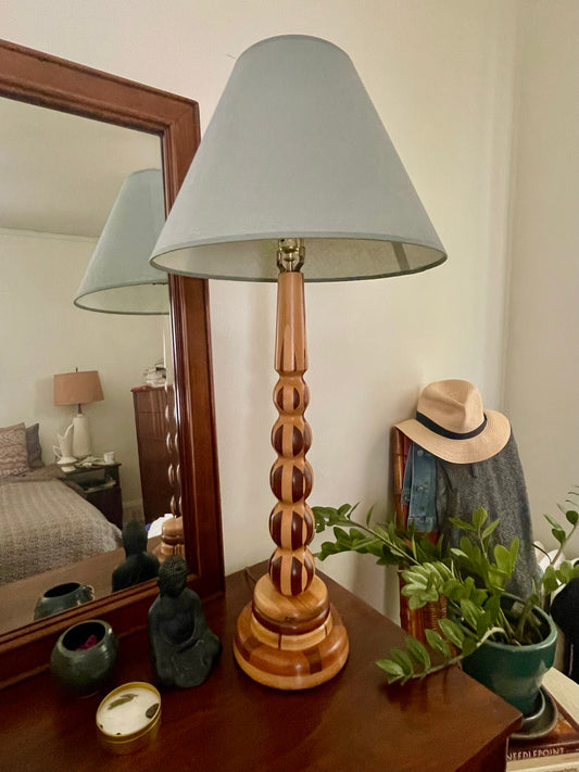 A wooden table lamp with a light blue-gray tapered linen shade and a turned parquetry wooden base, styled on a dark wood dresser in a bedroom setting with a mirror, hat, and plant.