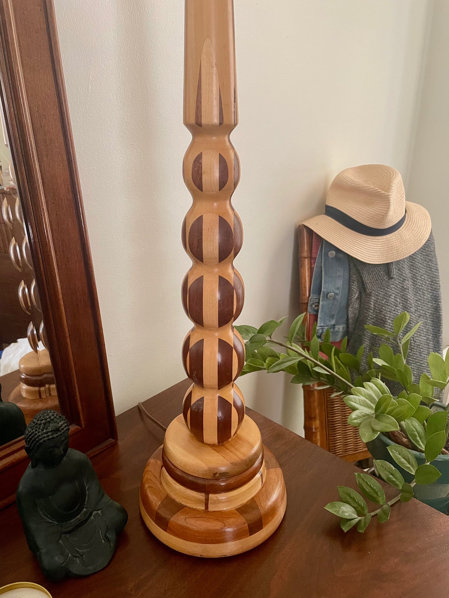 A wooden table lamp with a  turned parquetry wooden base, styled on a dark wood dresser in a bedroom setting with a mirror, hat, and plant.