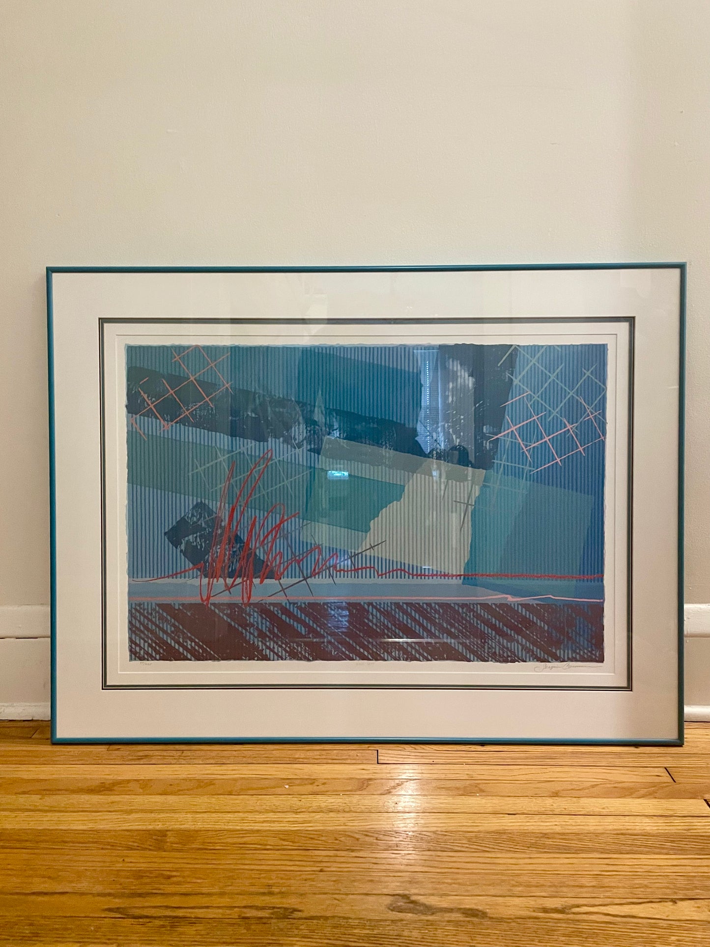 A framed abstract art print leaning against a beige wall on a wooden floor. The artwork features a complex composition of blue, teal, red, pink and mauve geometric patterns, enclosed in a white mat and a dark teal frame.