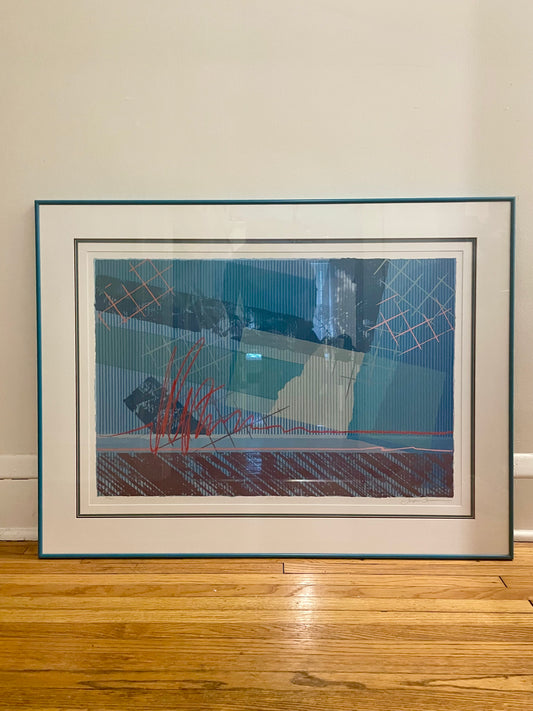 A framed abstract art print leaning against a beige wall on a wooden floor. The artwork features a complex composition of blue, teal, red, pink and mauve geometric patterns, enclosed in a white mat and a dark teal frame.