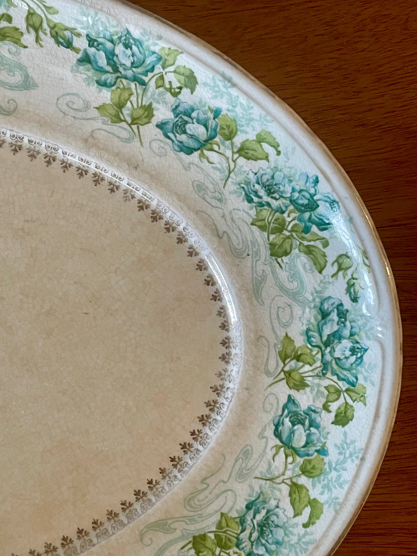 A close-up of an off-white porcelain serving platter, decorated with a border of blue roses, green leaves, and delicate scrollwork, set on a wooden surface.