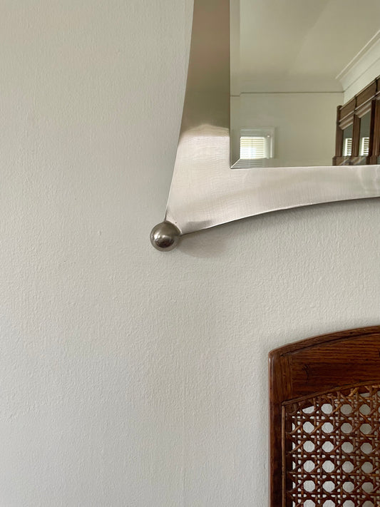 A close-up of a wall-mounted mirror with a silver metal frame, shown against a textured off-white wall with a wooden chair partially visible in the foreground.