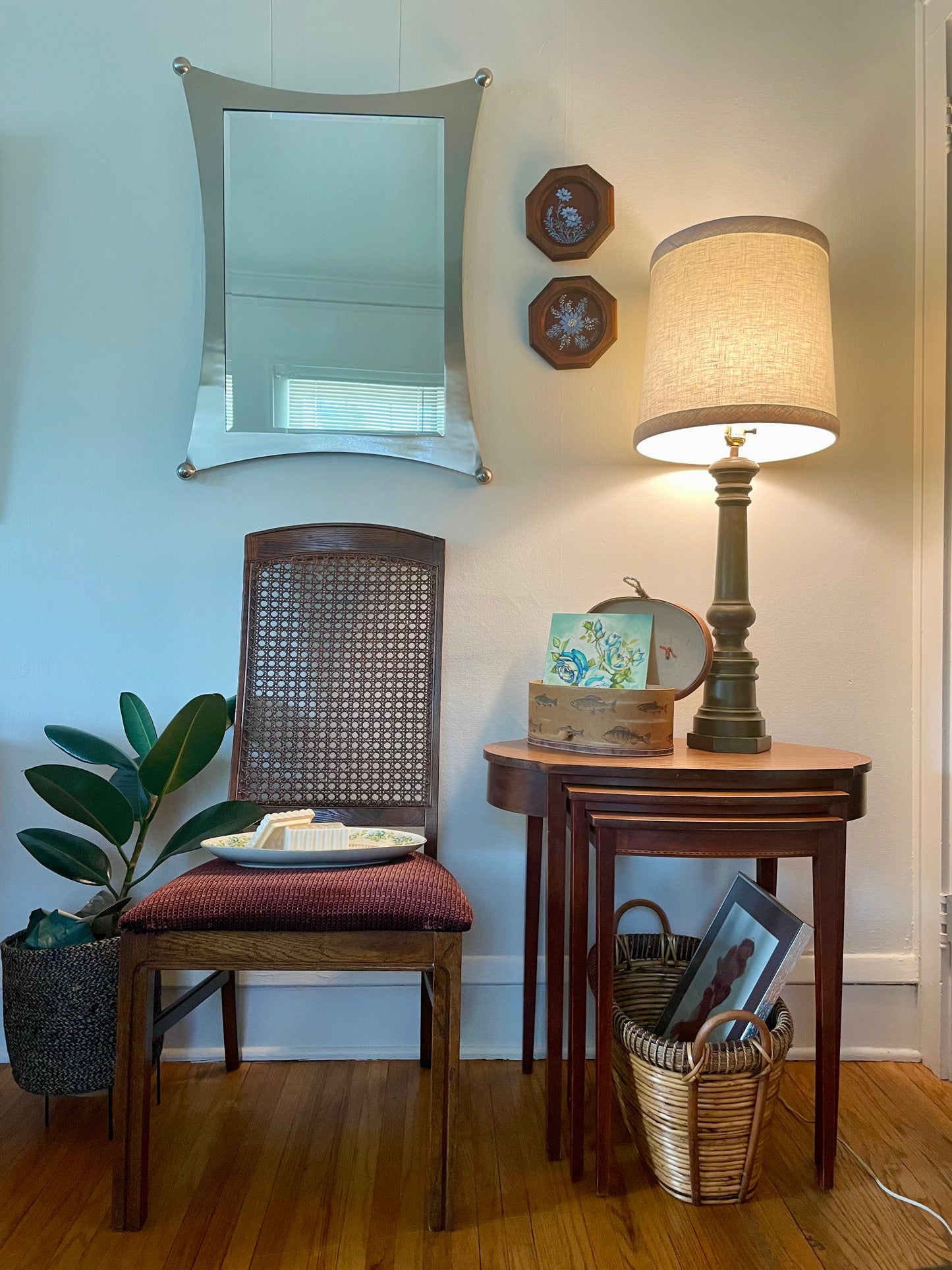 A styled interior corner featuring a wooden chair with a woven cane back and a red upholstered seat cushion, next to a small wooden side table holding a lit table lamp, a plant, and a silver mirror and other decorative items against a white wall.