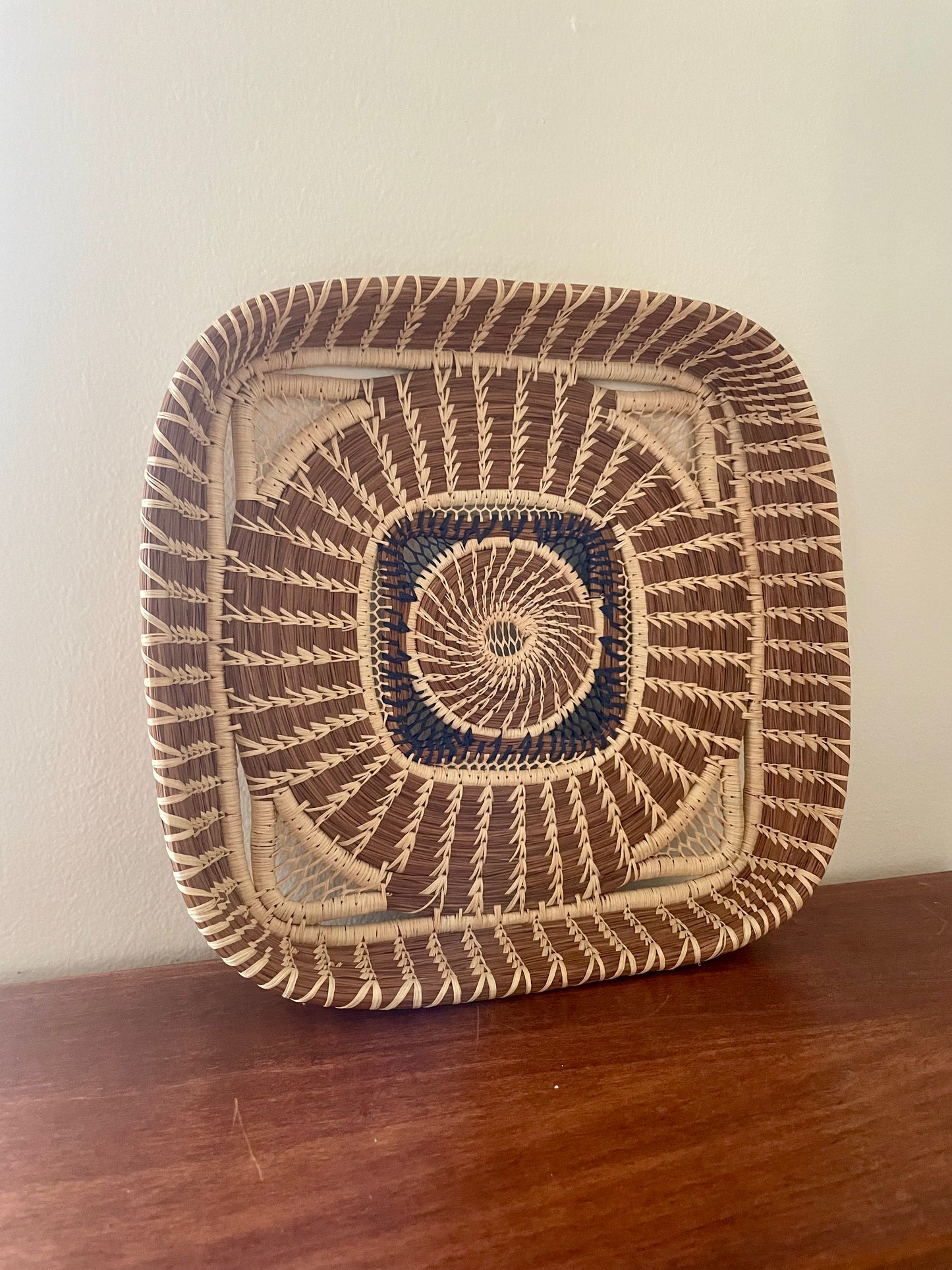 A square wild grass and pine needle woven basket with rounded corners, featuring a geometric pattern in dark brown, beige, and navy blue, resting on a wooden surface against a plain wall.