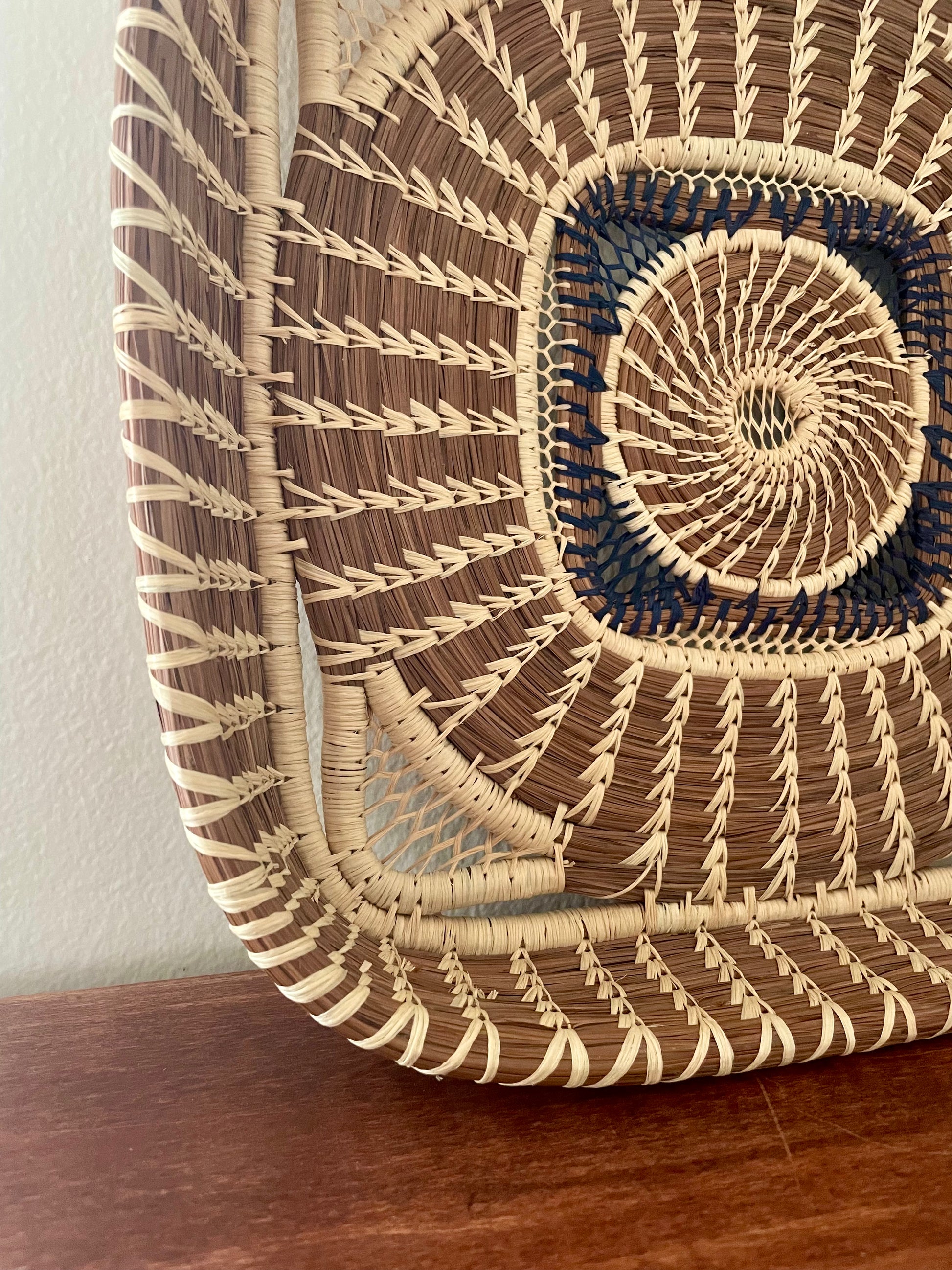 A close-up of a hand-woven wild grass and pine needle basket or tray with a geometric spiral pattern in natural beige, medium brown, and dark navy blue, resting on a wooden surface against a light wall.