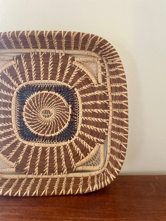 A square wild grass and pine needle woven basket with rounded corners, featuring a geometric pattern in natural beige, medium brown, and a central blue accent, resting on a dark wooden surface against a plain off-white wall.