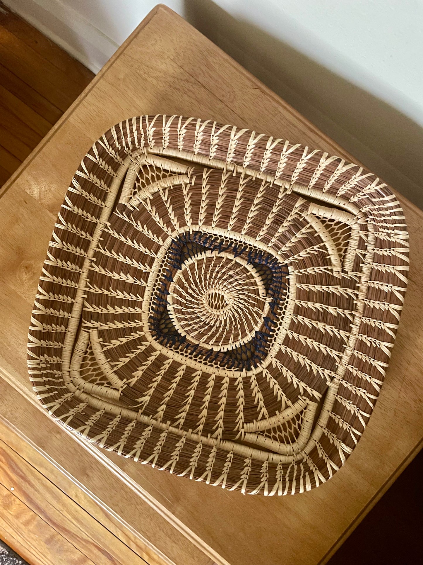 A square basket with a shallow raised rim featuring a light woven spiral pattern in the center surrounded by navy weaving, and then interwoven light and dark wild grass and pine details, shown from above on a wooden table.