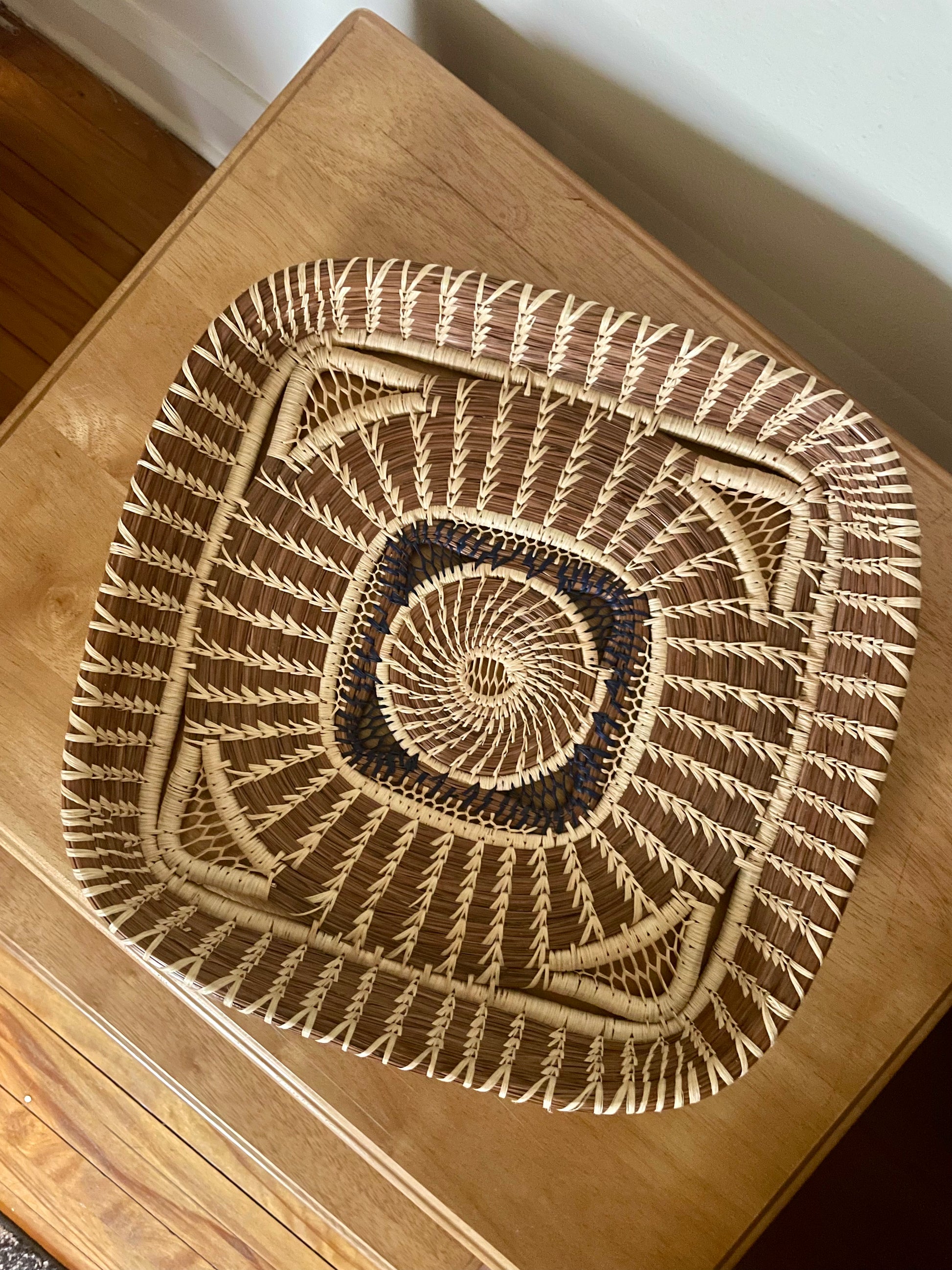 A square basket with a shallow raised rim featuring a light woven spiral pattern in the center surrounded by navy weaving, and then interwoven light and dark wild grass and pine details, shown from above on a wooden table.