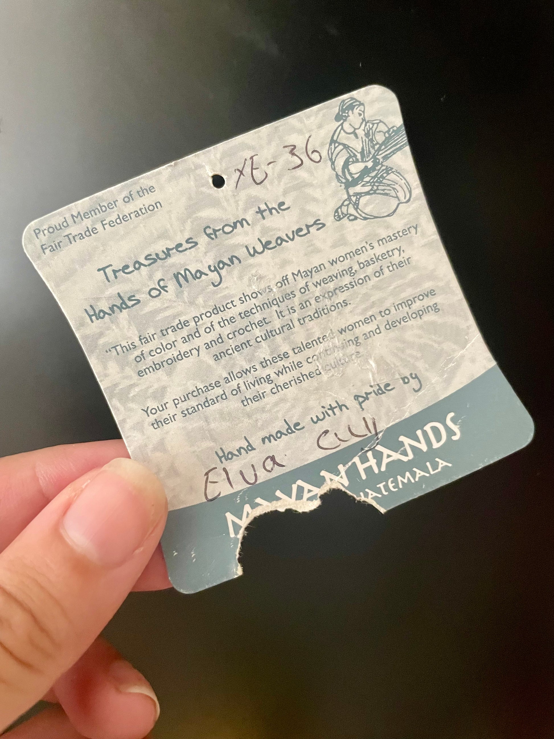 A hand holding a paper hangtag for a product from Mayan Hands Guatemala, member of the Fair Trade Federation and reading “Handmade with pride by Elva Cuy”.