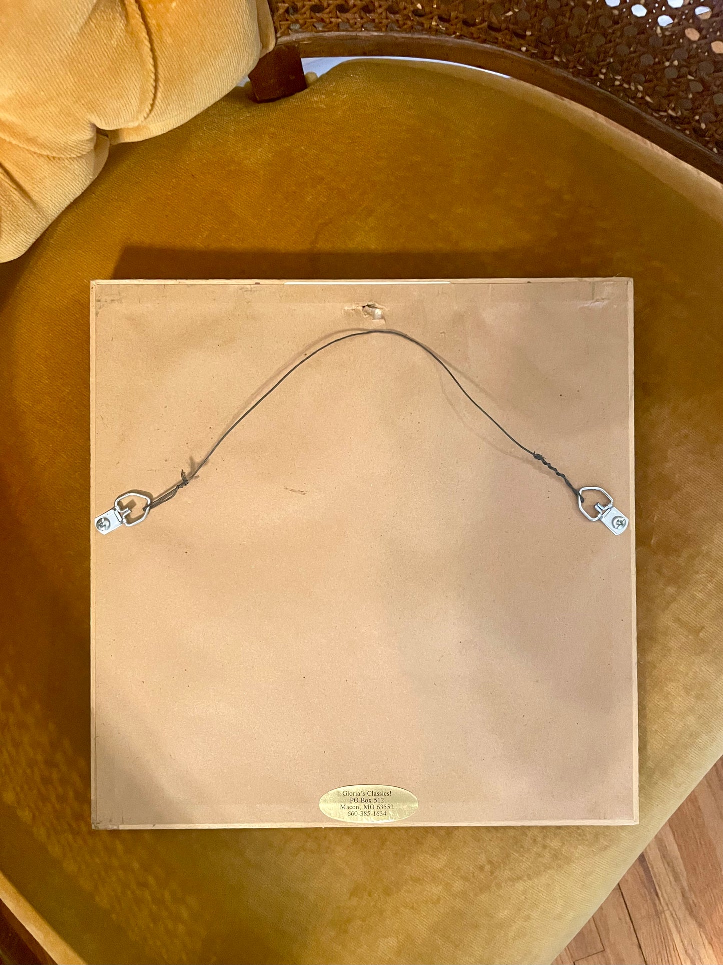 The back of a square frame, showing a tan backing board, hanging wire, and two metal hangers, placed on a mustard yellow upholstered chair.