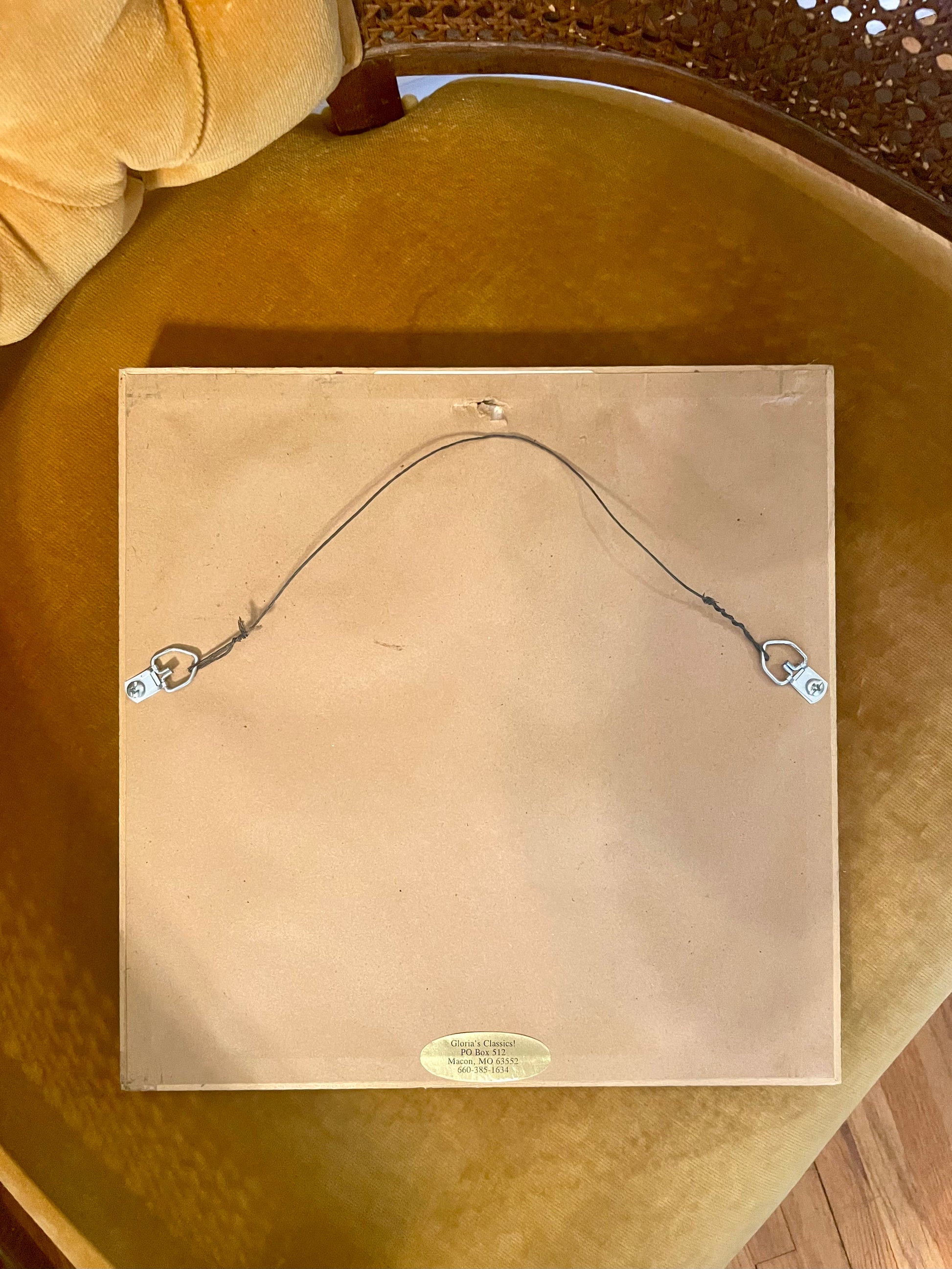 The back of a square frame, showing a tan backing board, hanging wire, and two metal hangers, placed on a mustard yellow upholstered chair.
