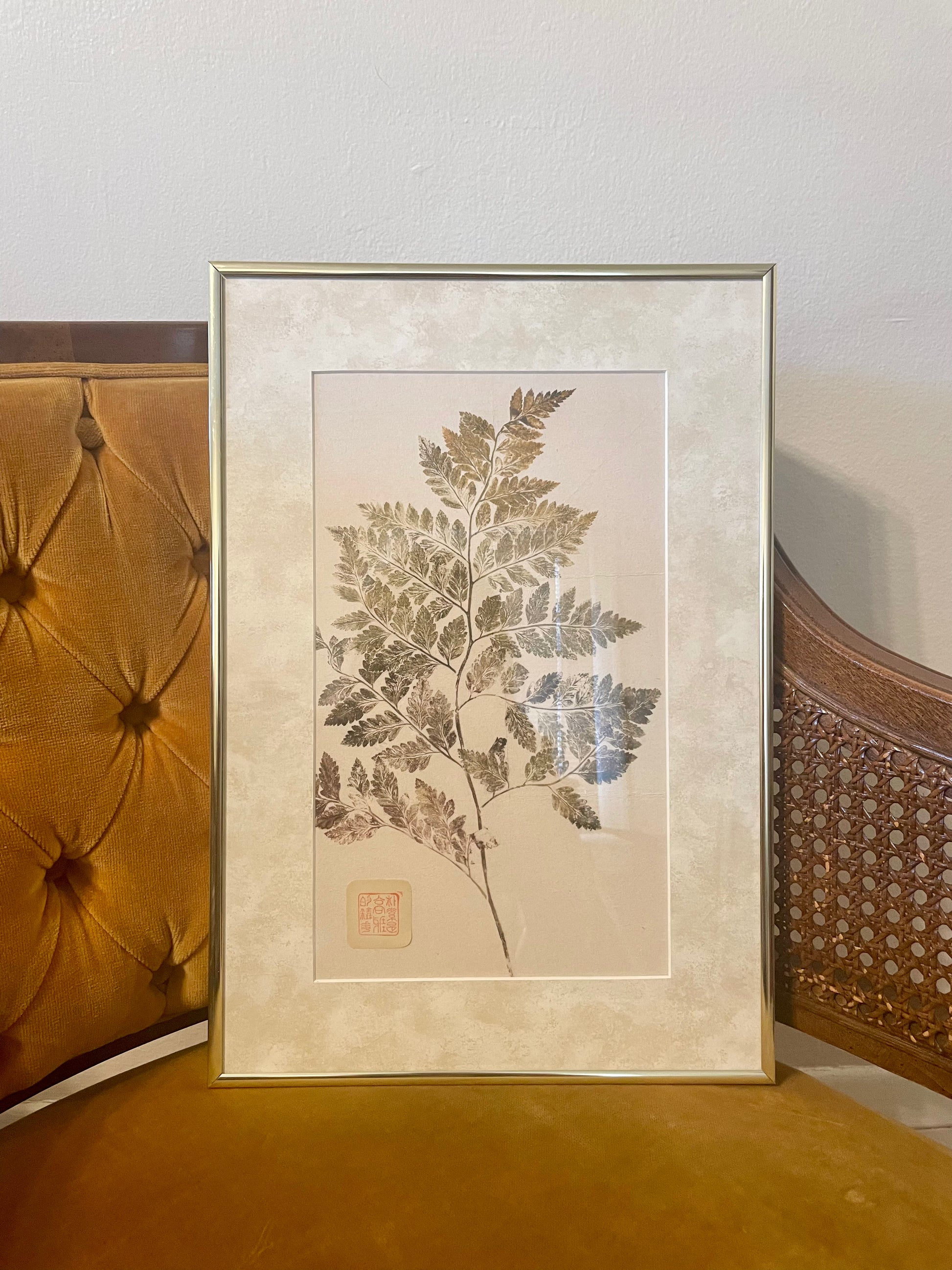 A framed botanical nature print featuring a detailed fern, styled leaning against a tufted mustard yellow velvet chair.