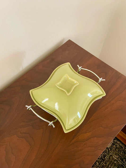 An avocado green ceramic wavy square serving dish with a matching lid, featuring chrome handles on a dark wood table.