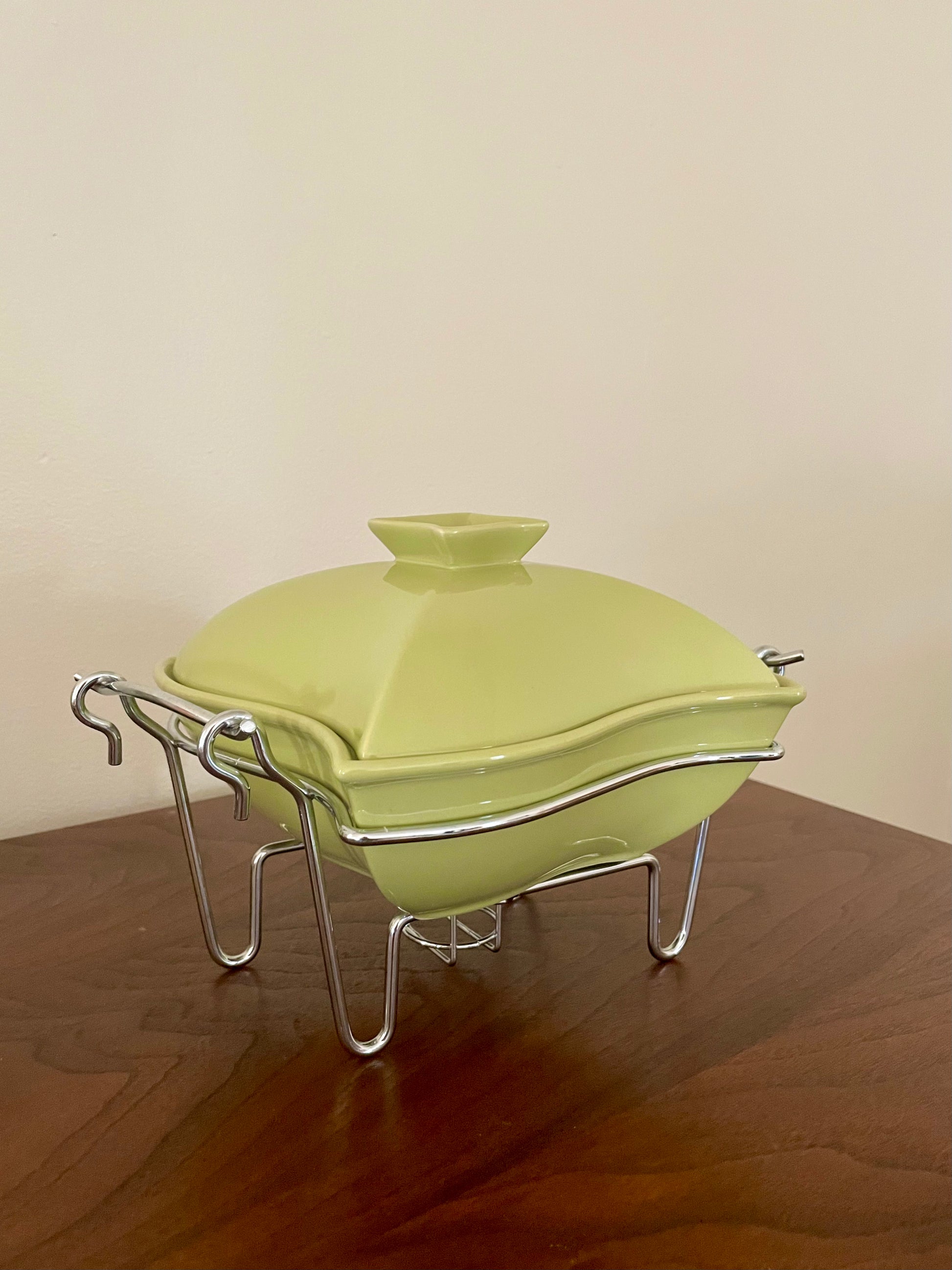 An avocado green ceramic chafing dish with a matching lid, resting on a silver metal wire stand on a wooden table against a plain beige wall.