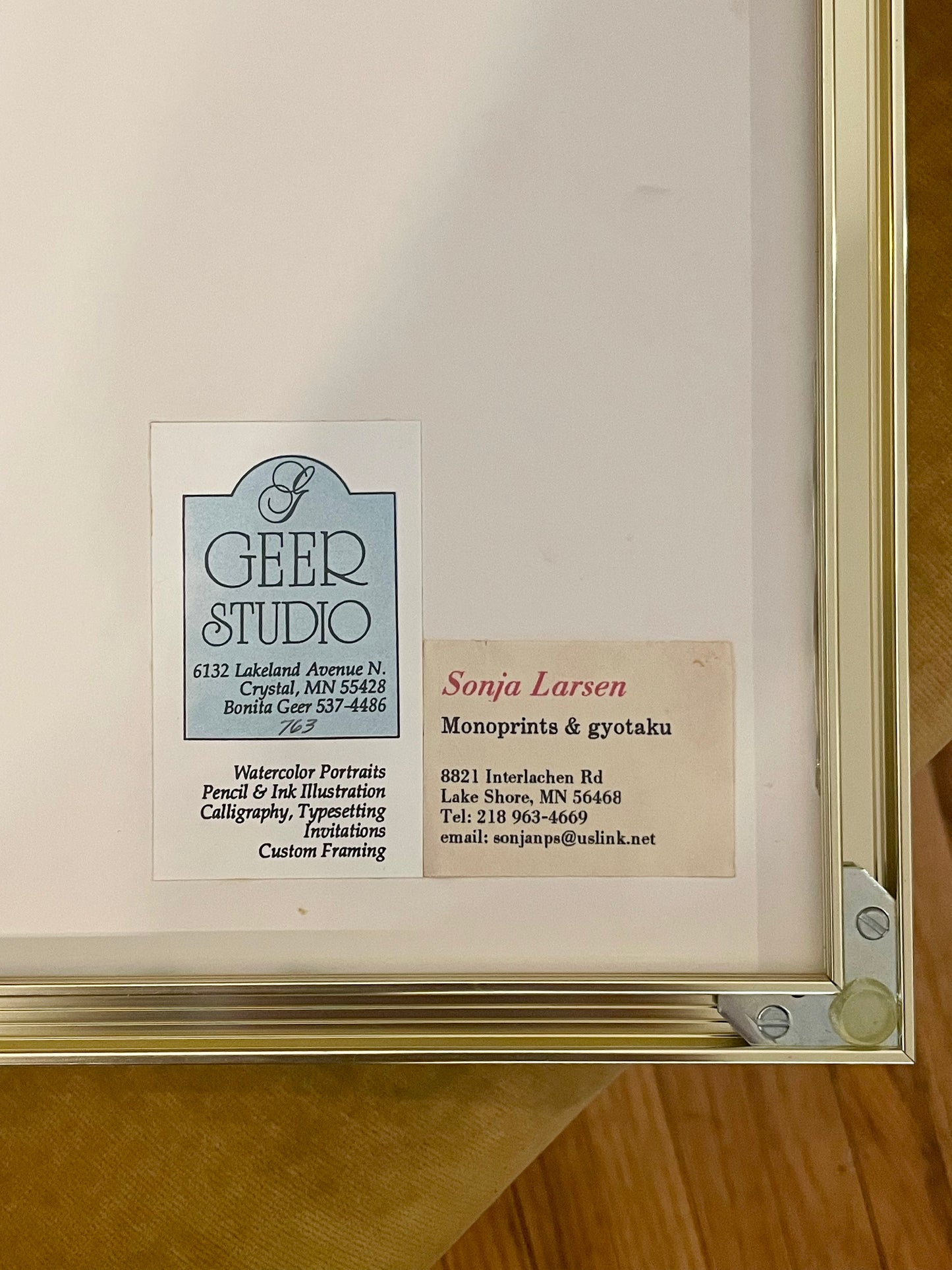 Backside of a framed artwork with a gold-toned frame and white backing, featuring the framer's 'Geer Studio' business card and the artist Sonja Larsen’s card.