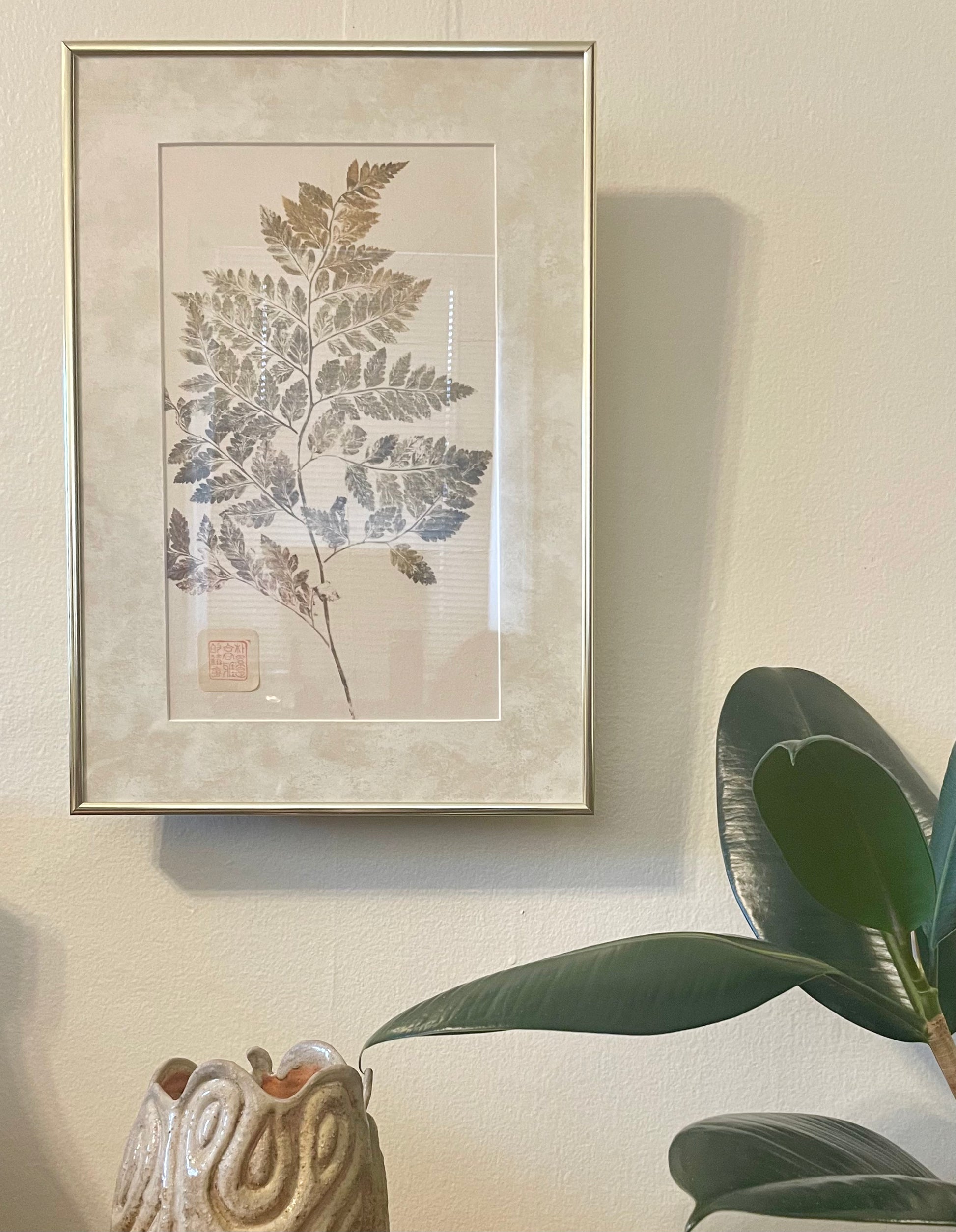 A framed botanical art print featuring a pressed fern leaf, styled on a beige wall with a green plant and a ceramic vase in the foreground.