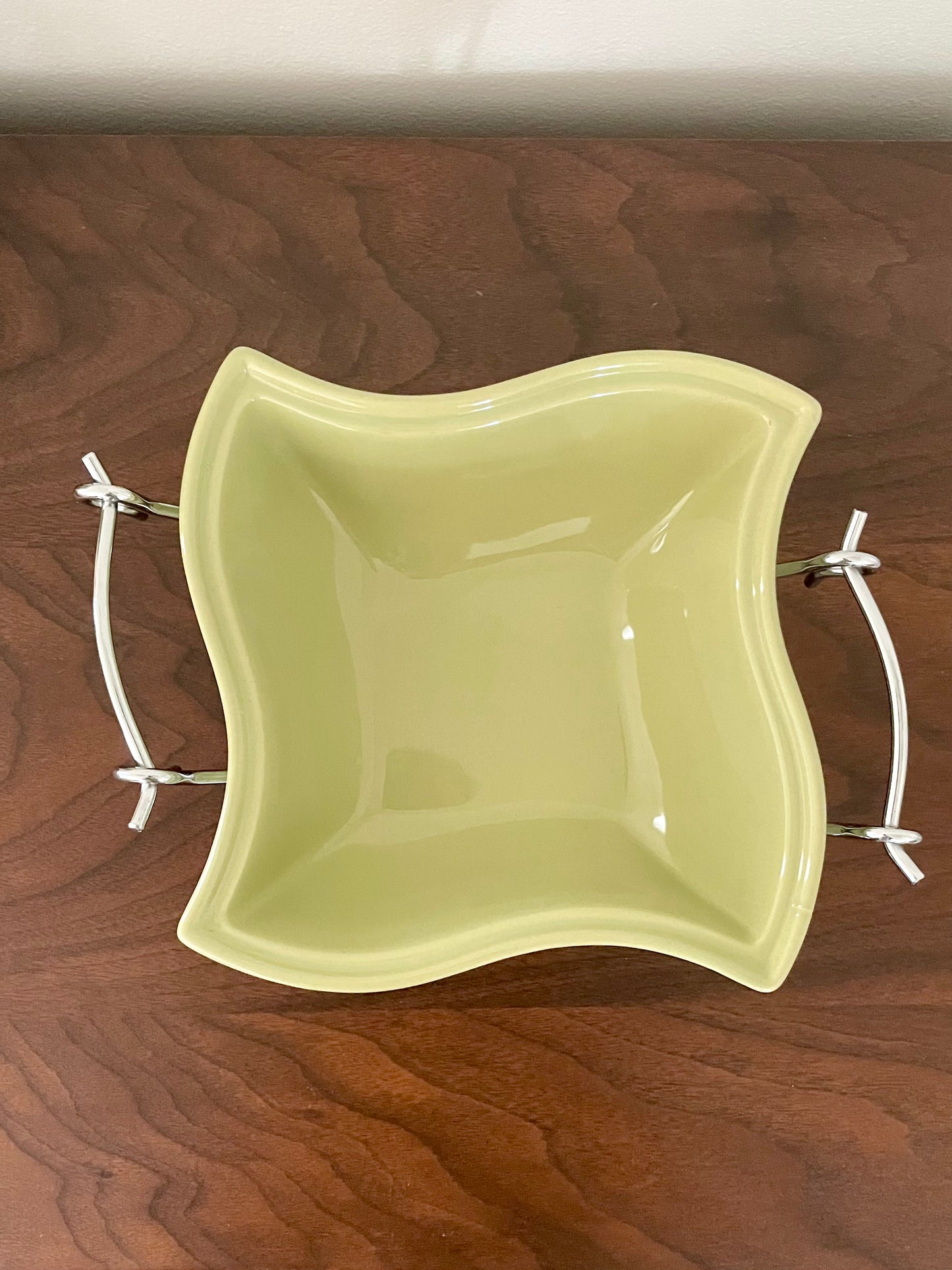 A square ceramic bowl with wavy edges in an avocado green glaze, featuring two chrome metal  handles, shown on a dark wood tabletop.