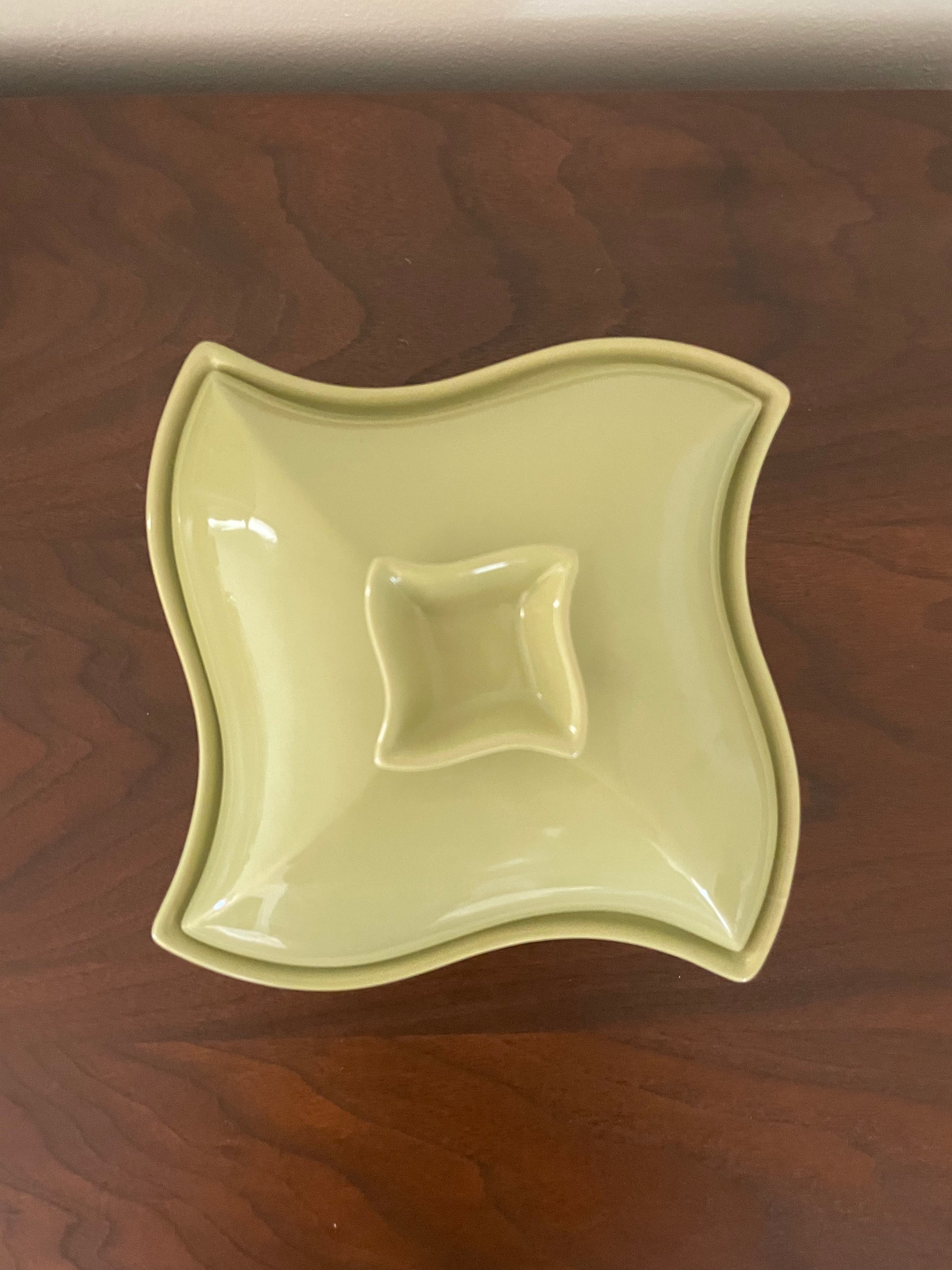 A square ceramic dish with wavy edges and a small central recess, shown from above on a dark wood surface. The dish is avocado green.