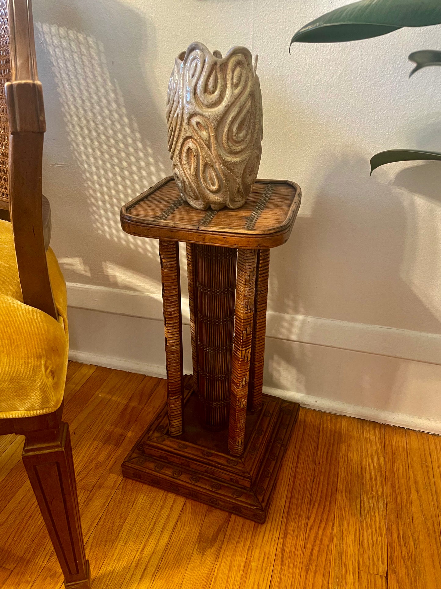 A carved wood and rattan-wrapped side table with a square top, four legs, and a center support, holding a textured beige ceramic vase, set in a room with hardwood floors, a white wall, and a yellow chair partially visible on the left.