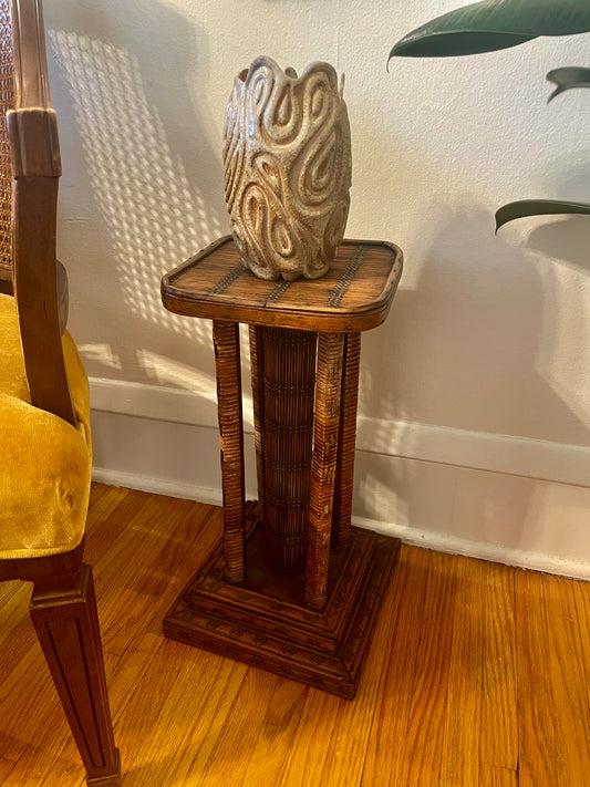 A carved wood and rattan-wrapped side table with a square top, four legs, and a center support, holding a textured beige ceramic vase, set in a room with hardwood floors, a white wall, and a yellow chair partially visible on the left.