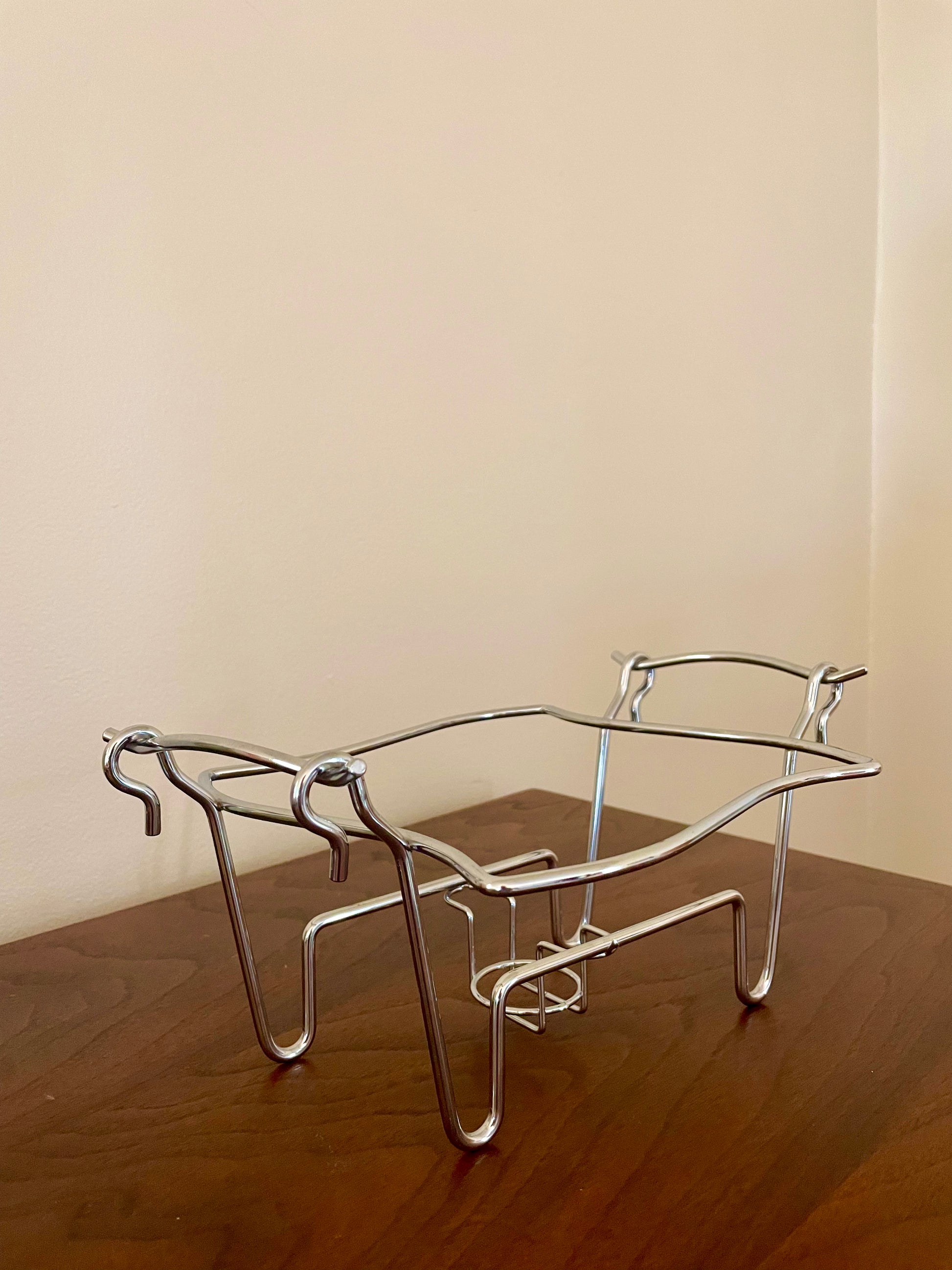 A chrome chafing dish stand resting on a dark wooden table against a plain beige wall.
