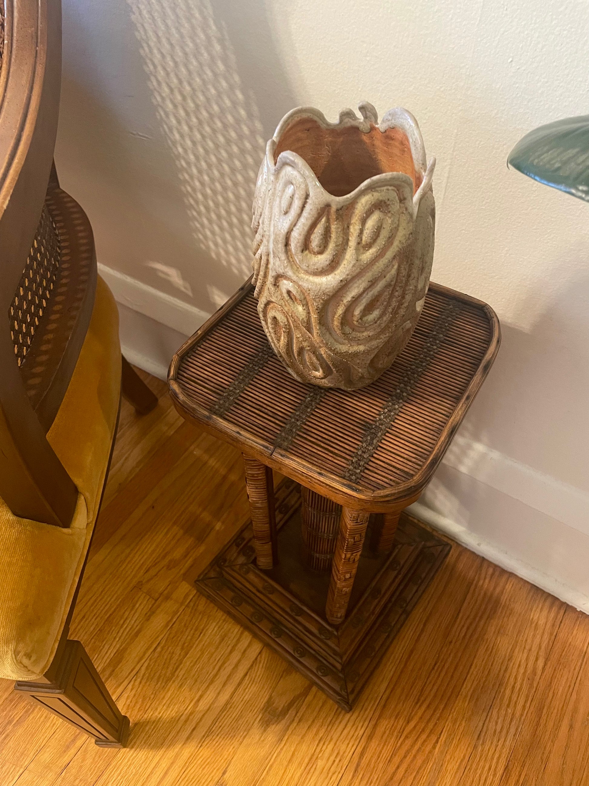 A textured, sculptural ceramic vase with a flared rim and swirling organic patterns, displayed on a small woven side table in a home setting.