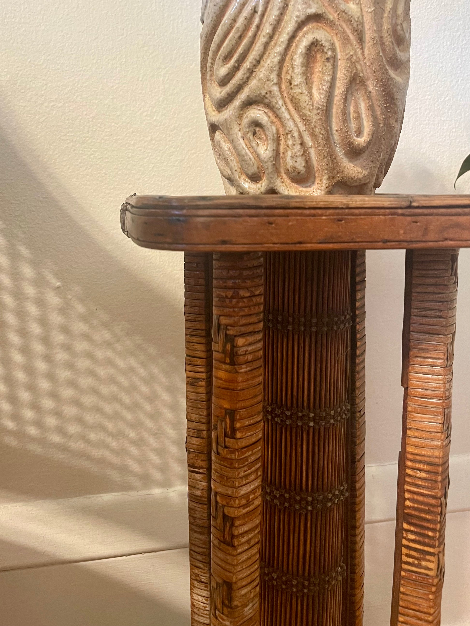 A close-up of a rattan and carved wood table with a carved ceramic vase sitting on top, set against a plain off-white wall with soft shadows.