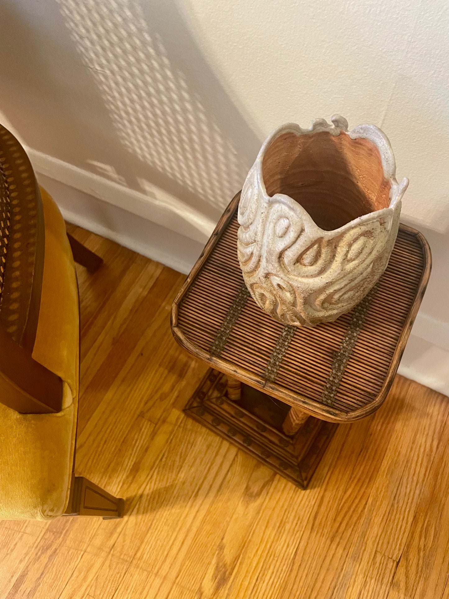 A textured, sculptural ceramic vase with a flared rim and swirling organic patterns, displayed on a small woven side table in a home setting.