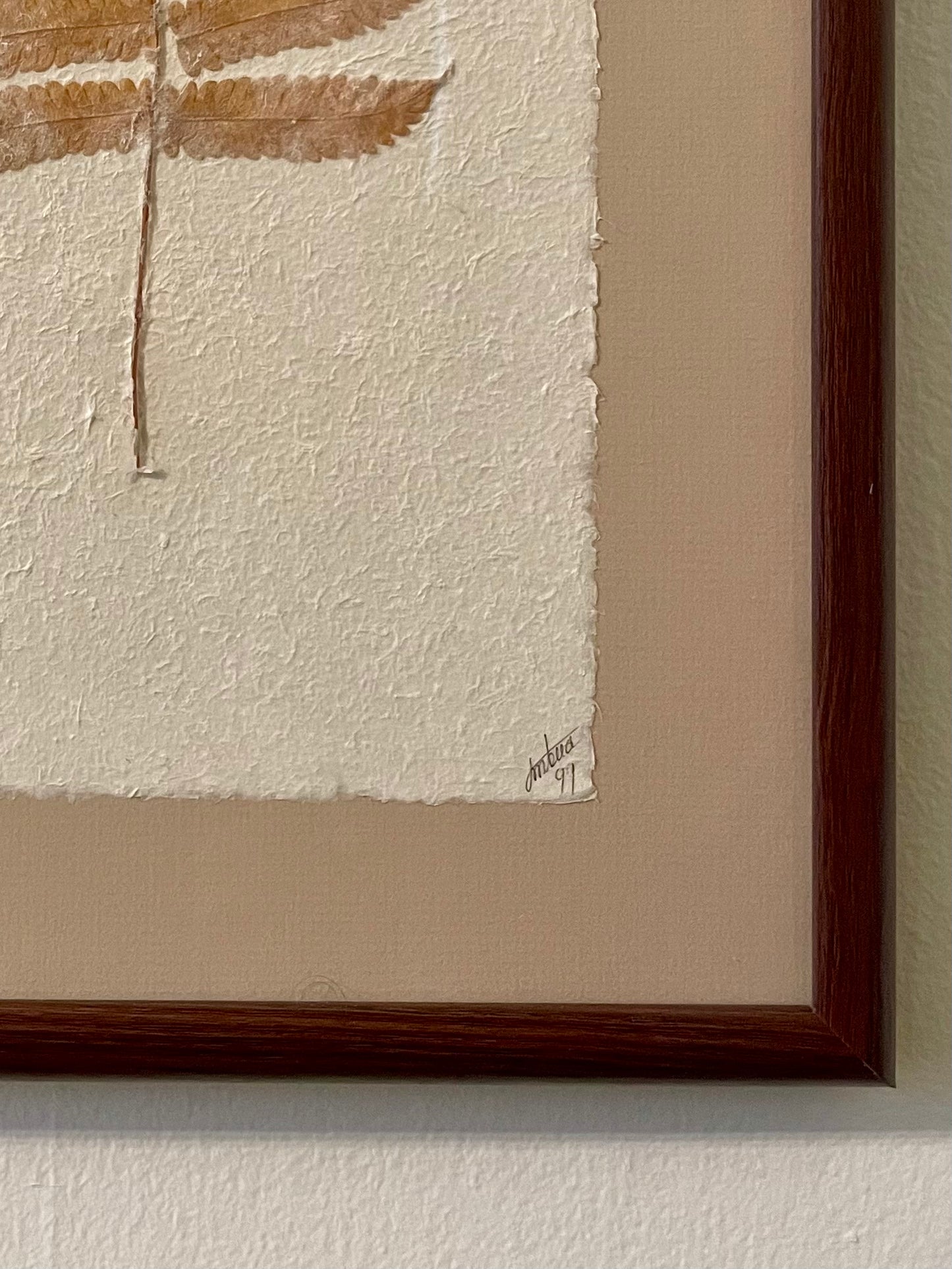 Close-up of a framed artwork showing a textured off-white paper panel with a partial view of a pressed fern, a beige mat, and a dark reddish-brown wooden frame mounted on a light wall. A signature and the date ’99 is written in script in the lower right corner.