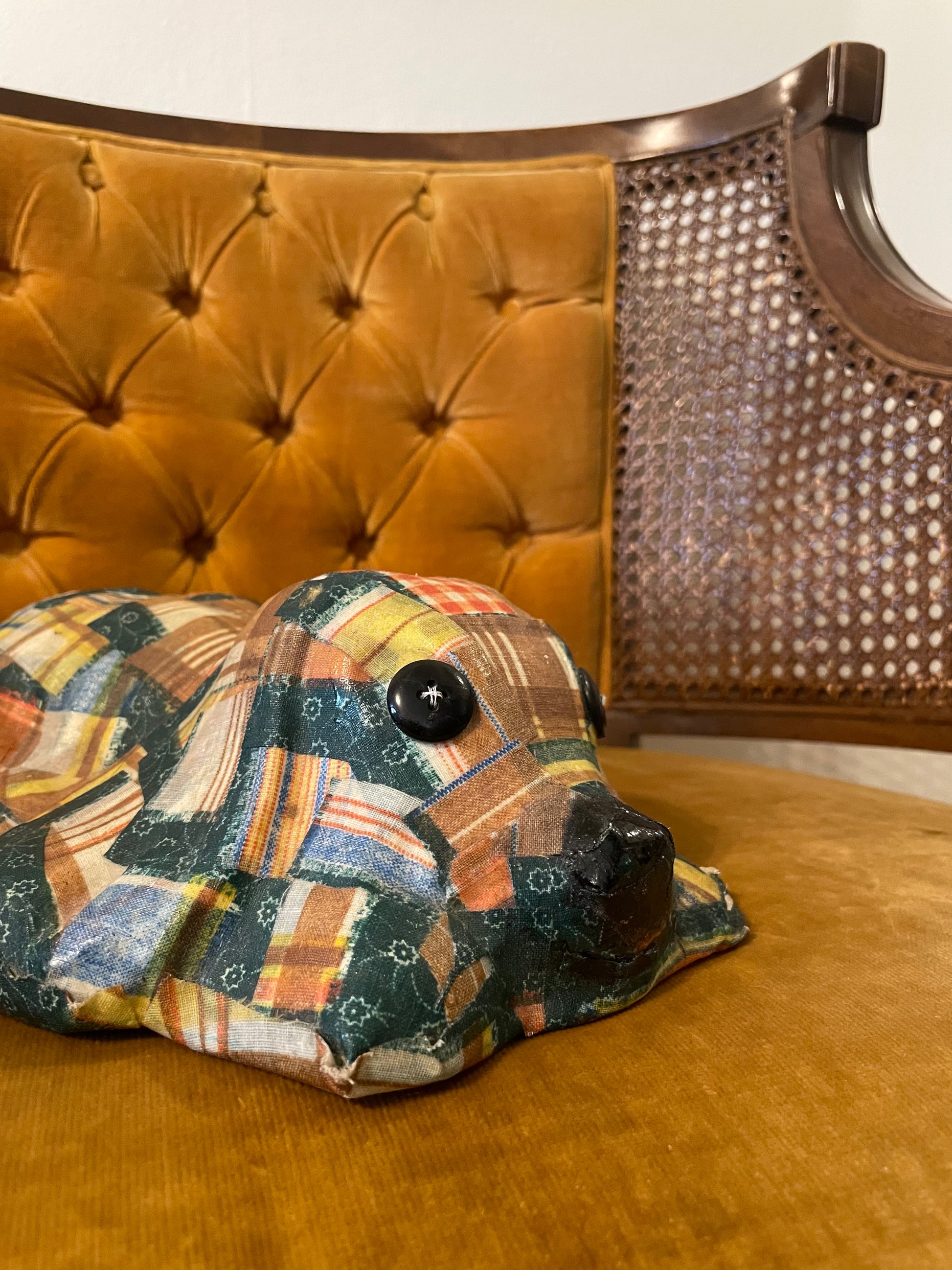A handmade fabric-covered dog sculpture with a multicolor patchwork pattern, featuring black button eyes and a black nose, resting on a mustard yellow velvet chair with a tufted back.