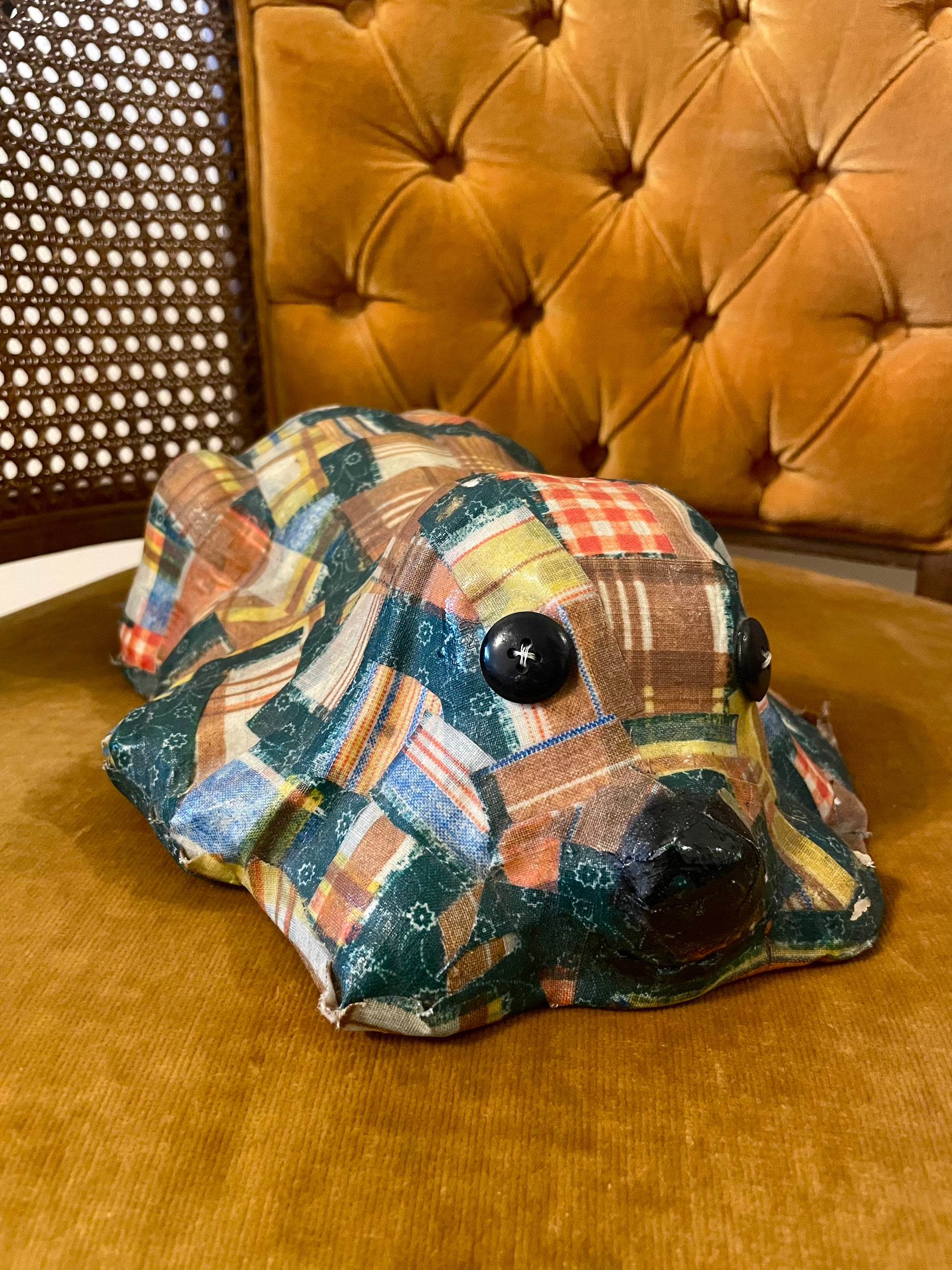 A handmade fabric-covered dog sculpture with a multicolor patchwork pattern, featuring black button eyes and a black nose, resting on a mustard yellow velvet chair with a tufted back.