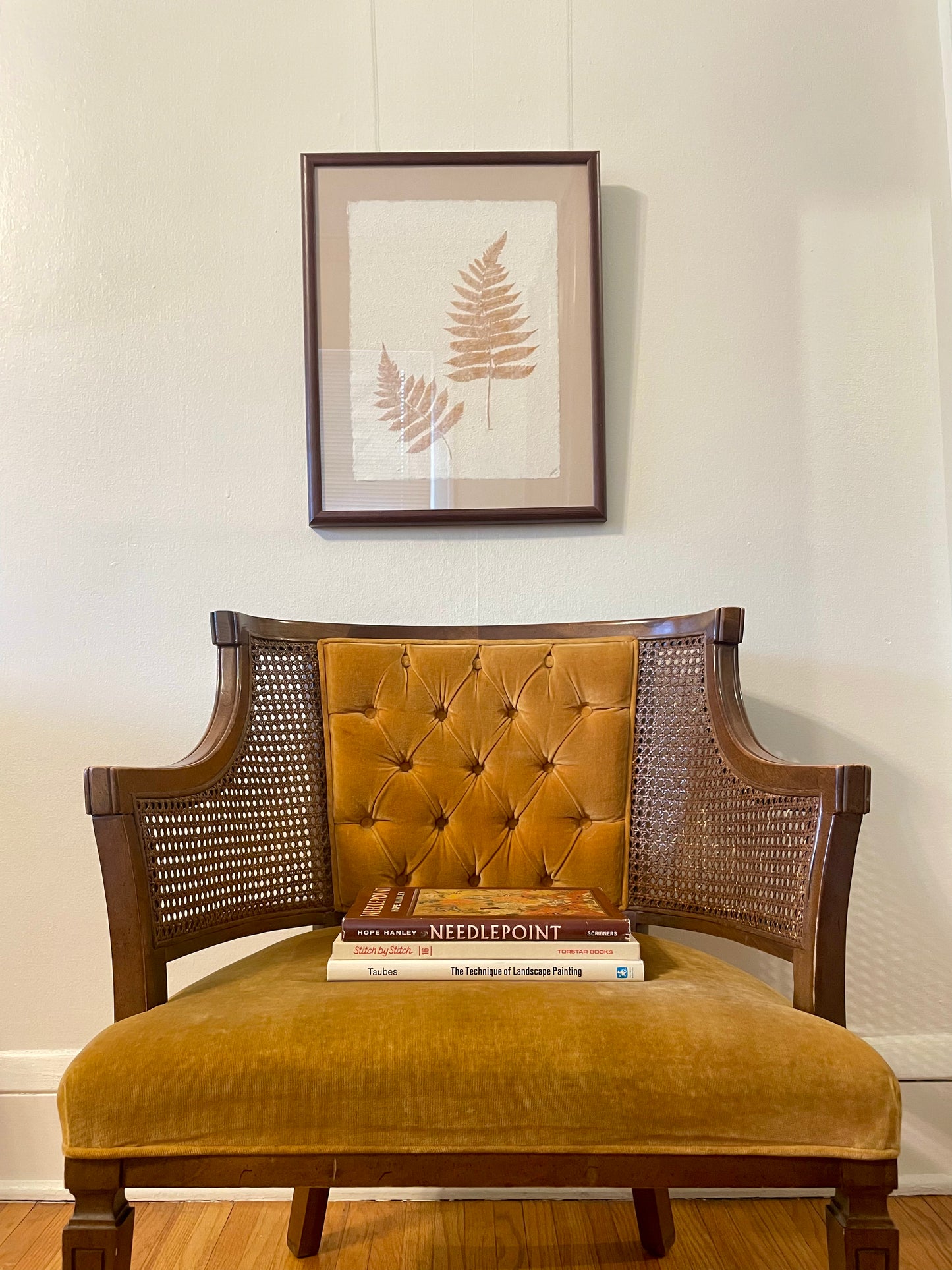 A vintage armchair with a mustard yellow tufted velvet backrest and seat cushion, dark brown wooden frame, and woven cane side panels, shown on a hardwood floor against a white wall with framed botanical art above.