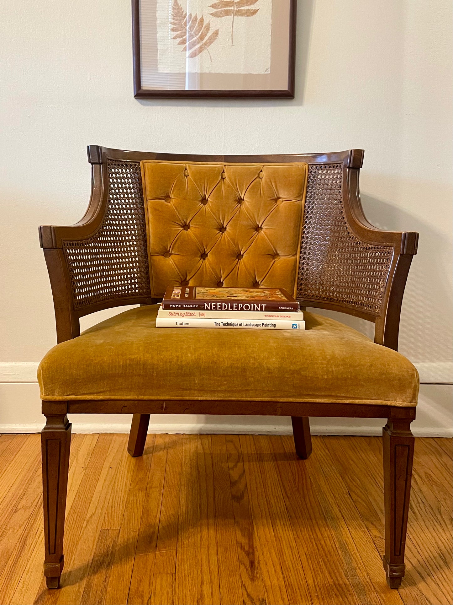 A vintage armchair with a mustard yellow tufted velvet backrest and seat cushion, dark brown wooden frame, and woven cane side panels, shown on a hardwood floor against a white wall with framed botanical art above.