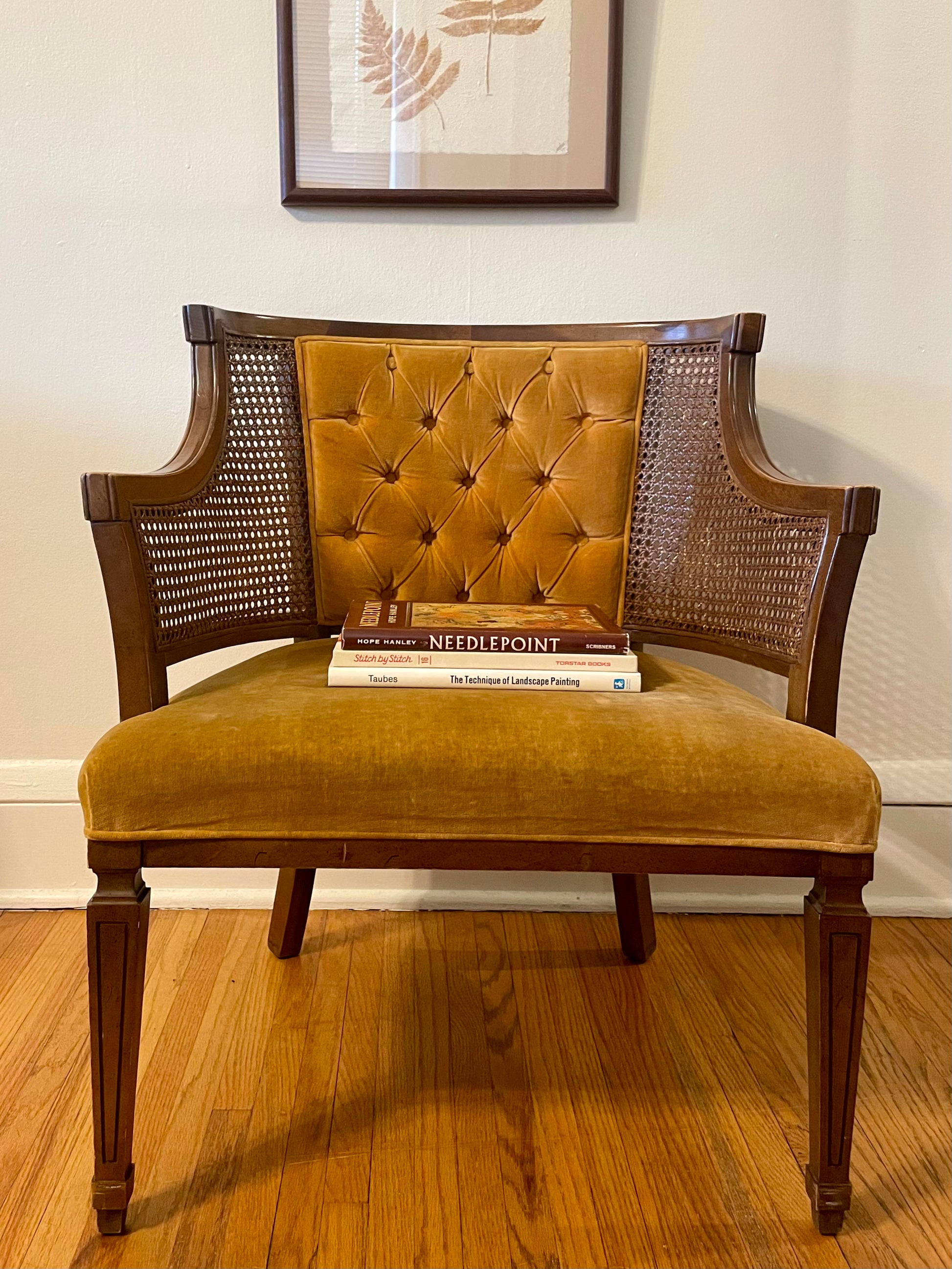 A vintage armchair with a mustard yellow tufted velvet backrest and seat cushion, dark brown wooden frame, and woven cane side panels, shown on a hardwood floor against a white wall with framed botanical art above.