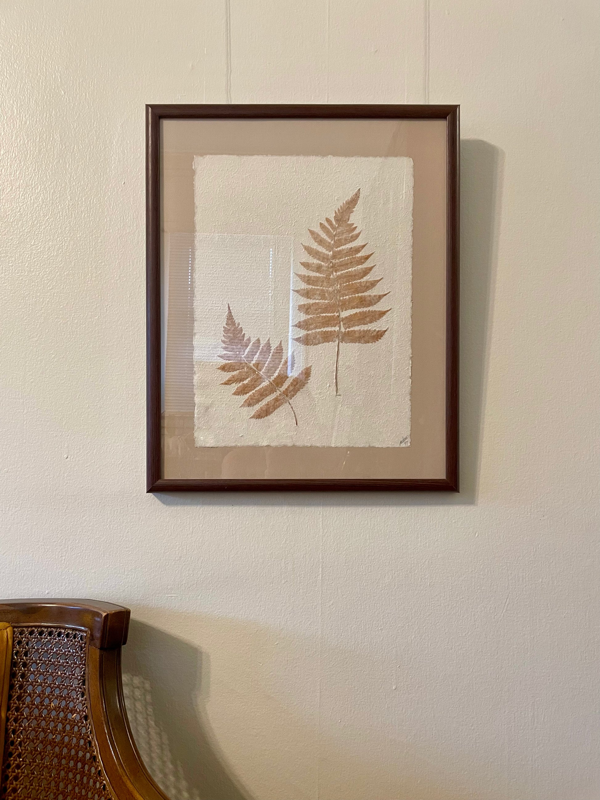 Framed botanical wall art featuring two pressed fern leaves on textured off-white paper, mounted with a beige mat and enclosed in a dark brown wooden frame, hanging on a cream-colored wall above a wooden chair.