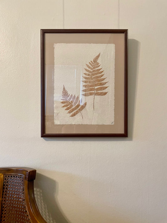 Framed botanical wall art featuring two pressed fern leaves on textured off-white paper, mounted with a beige mat and enclosed in a dark brown wooden frame, hanging on a cream-colored wall above a wooden chair.