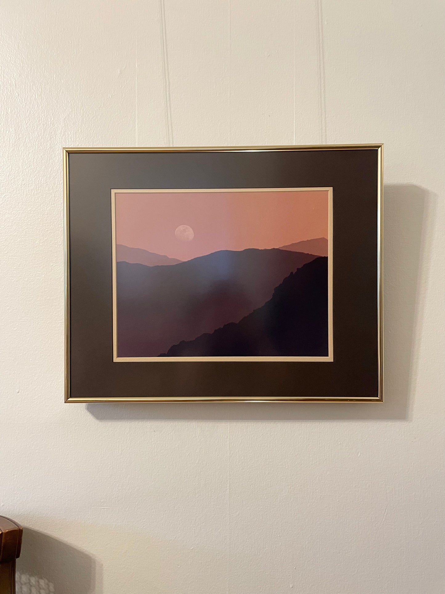 A framed landscape photograph depicting the silhouette of a mountain range under a pink and orange sky with a moon in the sky, mounted on a white wall.