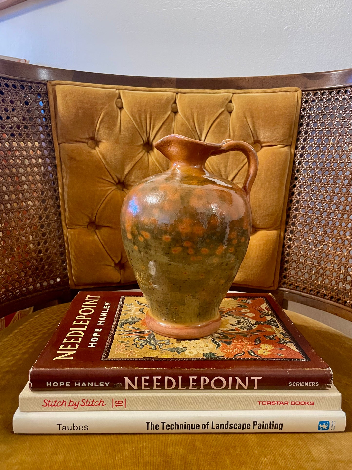 A handmade ceramic pitcher with a orange and green speckled glaze sits on a stack of three books in front of a tufted mustard-yellow chair.