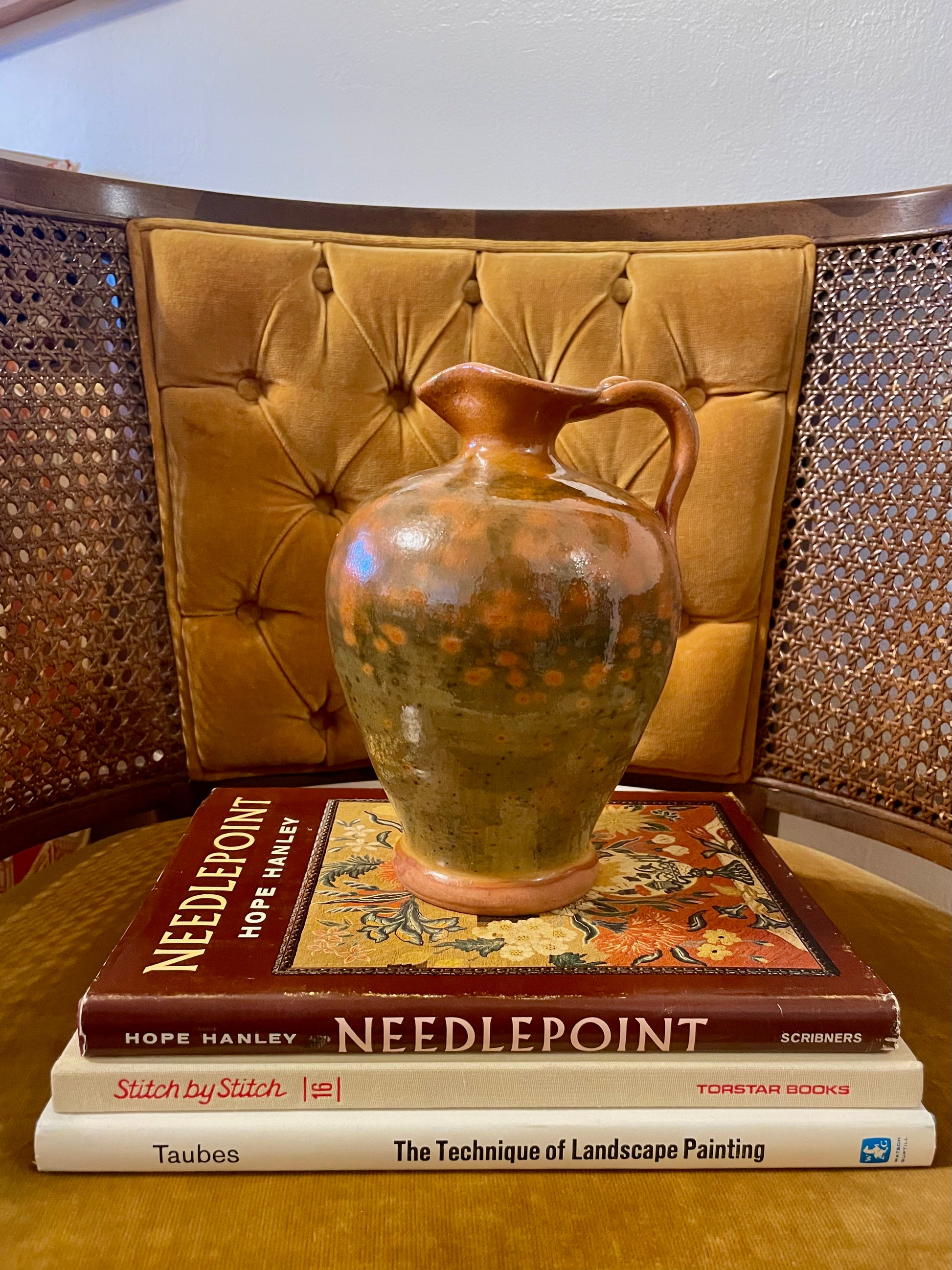 A handmade ceramic pitcher with a orange and green speckled glaze sits on a stack of three books in front of a tufted mustard-yellow chair.