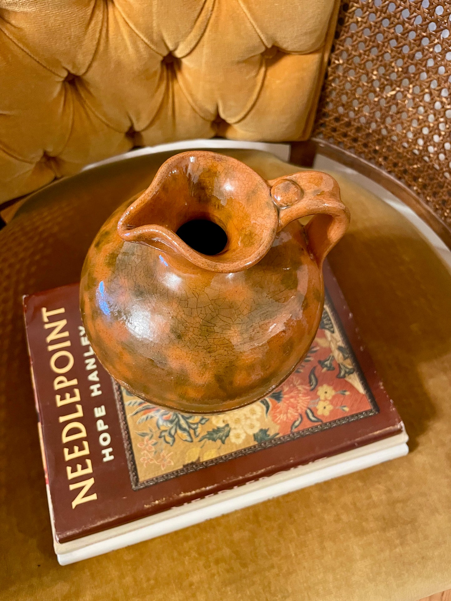 A handmade green and orange glazed ceramic pitcher with a handle, resting on a vintage book titled 'Needpoint' on a gold velvet chair.